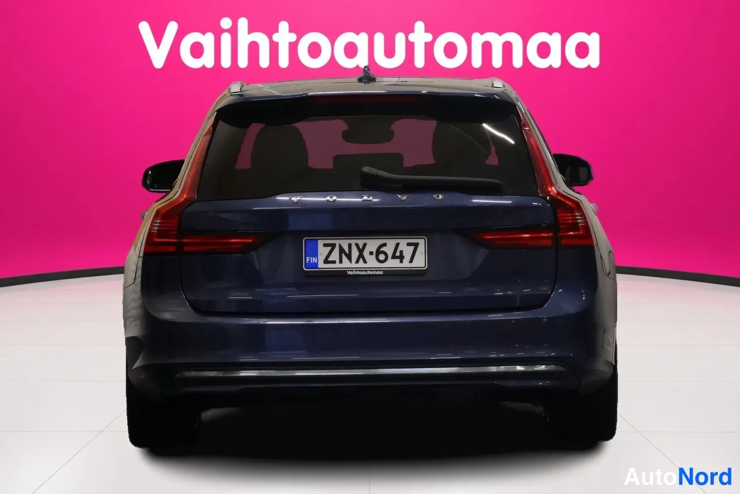 Takaa: Auton takaosa suoraan takaa, rekisteritunnus, tummansininen koripanelo vaaleanpunaista taustaa vasten.