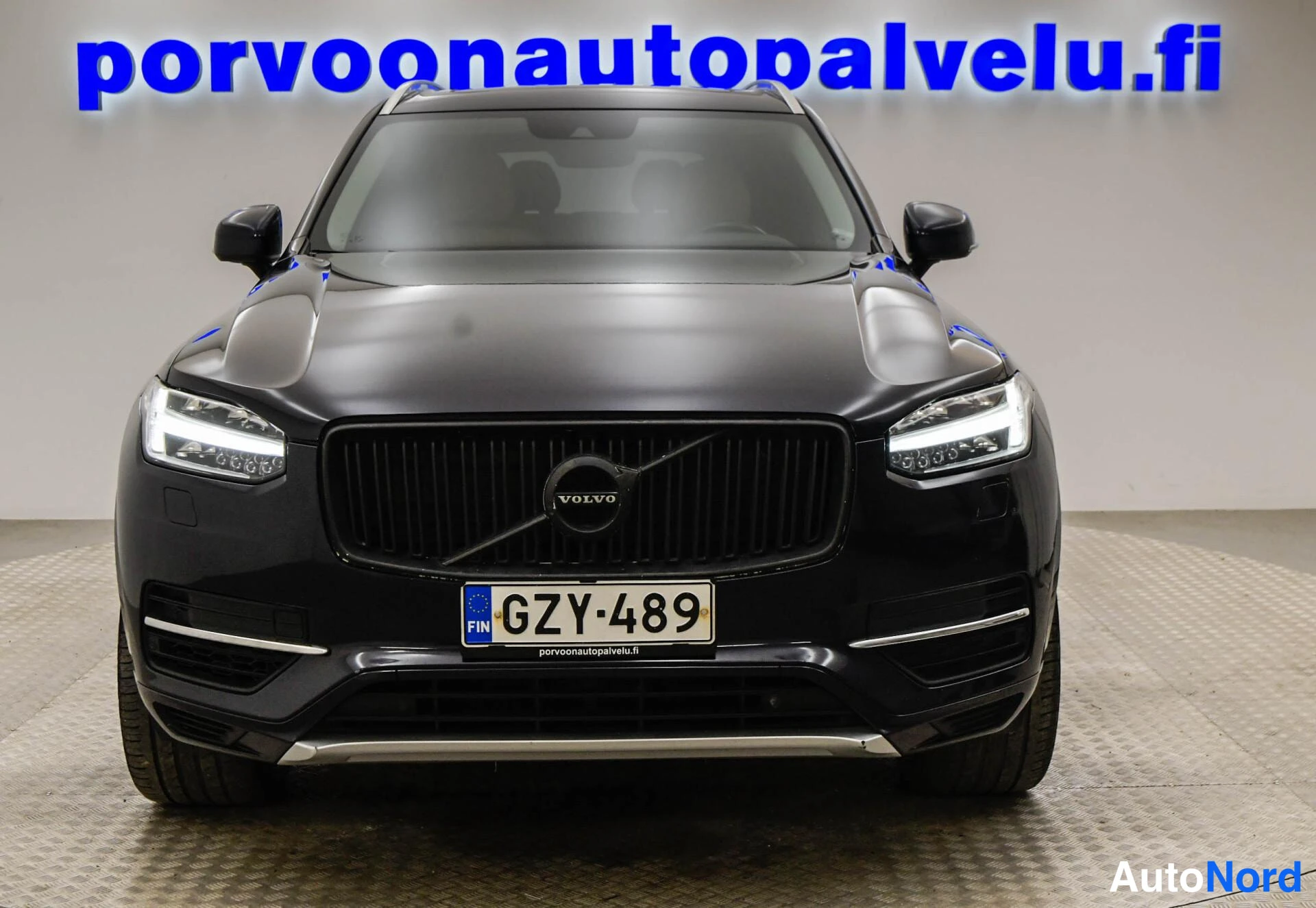 Edestä: Mustan tila-auton suora etunäkymä: Volvo-merkki keskellä säleikköä, -rekisterikilpi ja kromikoristeet puskurissa.