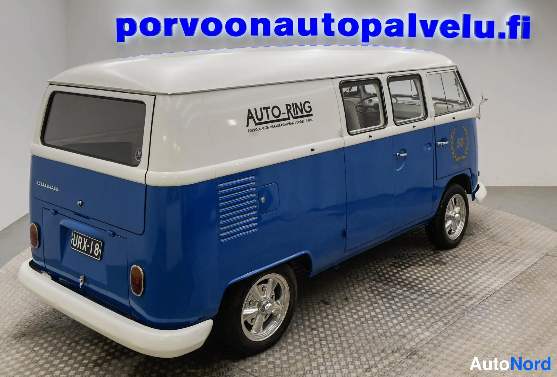 Takaviisto: Sinivalkoisen pakettiauton takakulma: takamoottorin ilmastointiritilät, rekisteritunnus ja sivulla Auto-Ring-teksti.