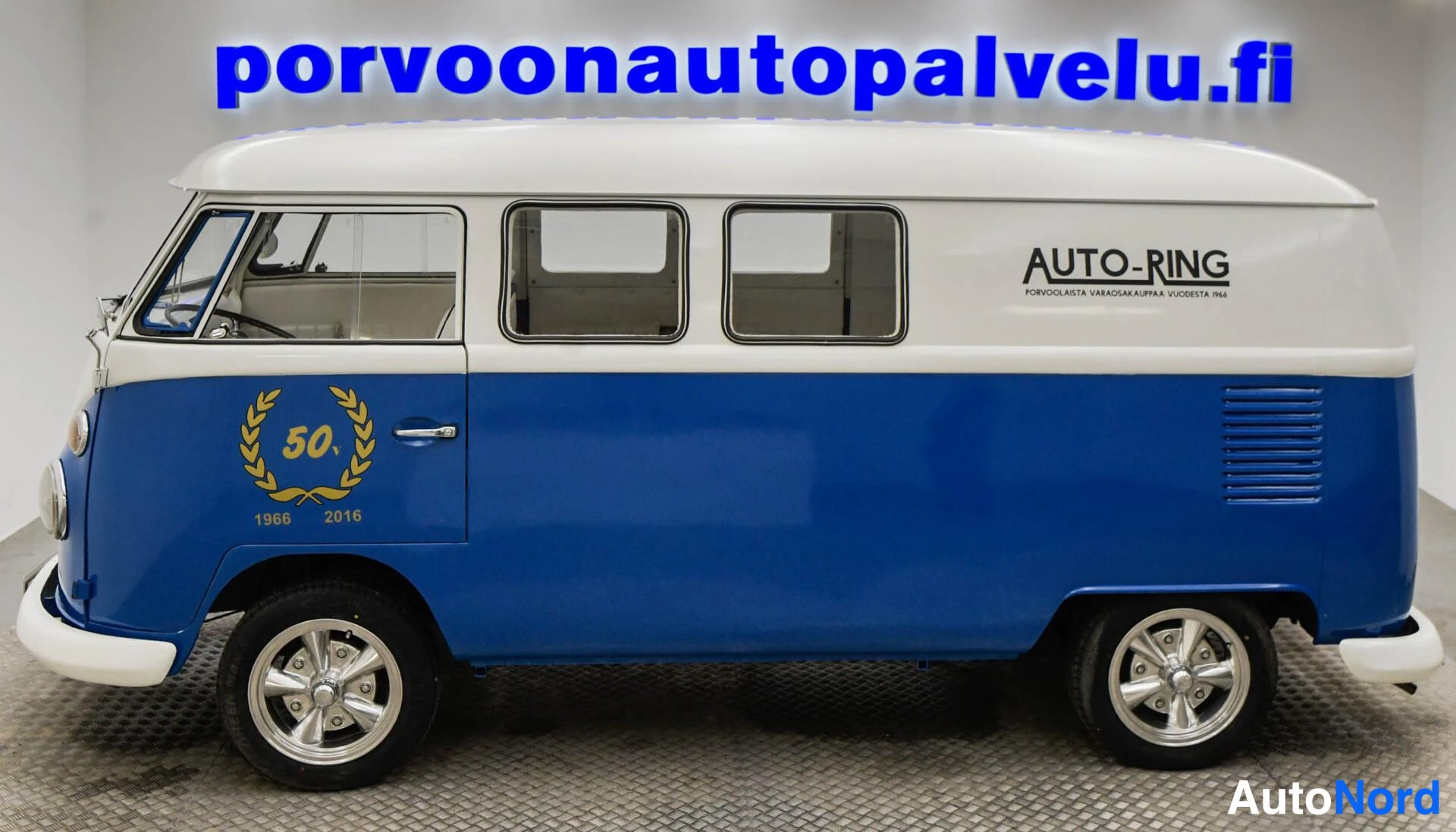 Etuviisto, nokka vasemmalle: Sinivalkoinen Volkswagen pakettiauto sivulta etukulmasta, kyljessä AUTO-RING-teksti ja kultainen 50-vuotis-embleemi vuosilla 1966–2016.