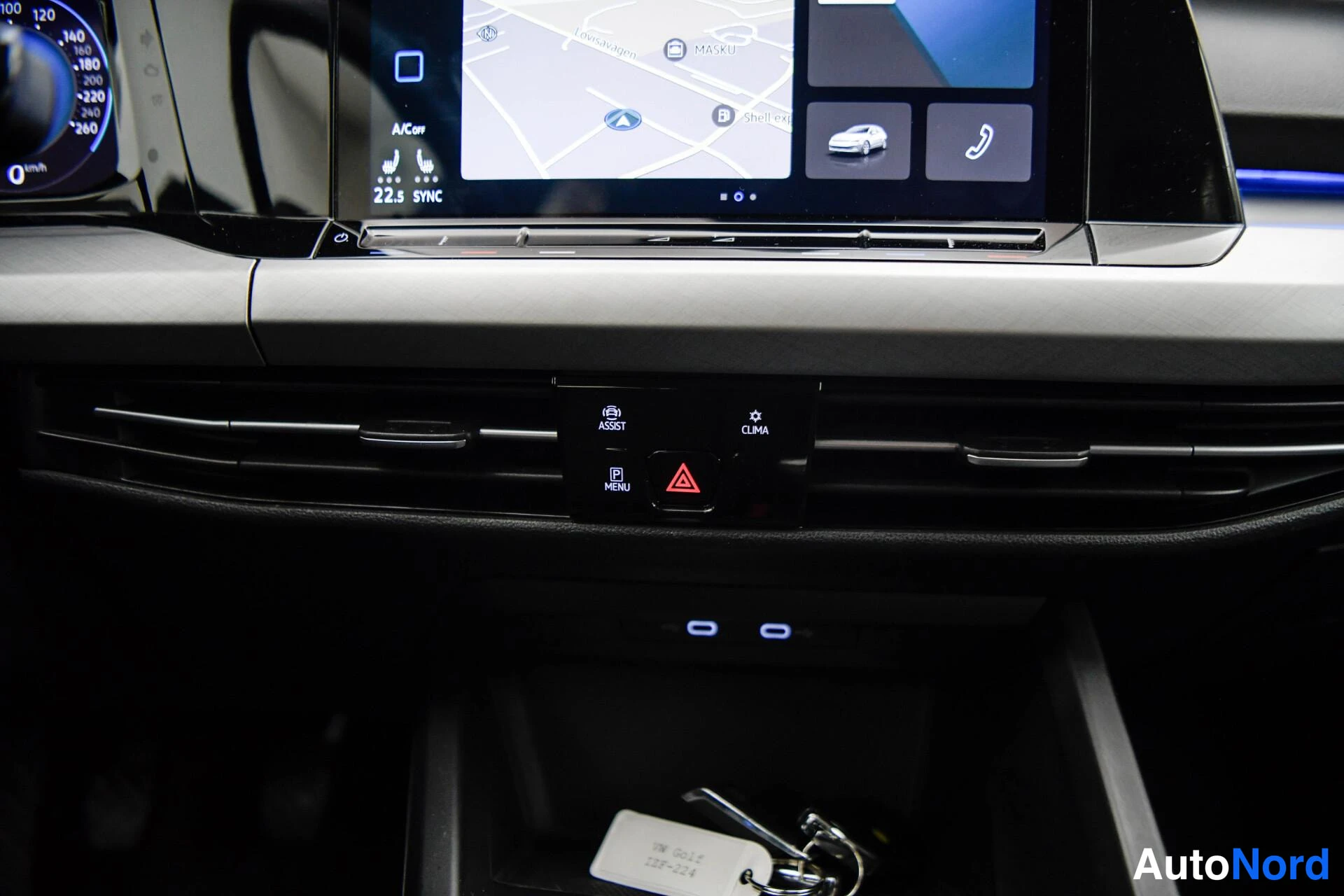 Yksityiskohta: Kojelaudan keskialue: infotainment-näyttö kartalla, AC/ON- ja SYNC-napit, CLIMA- ja ASSIST-toiminnot, varoitusvalo ja avaimet säilytyslokerossa.
