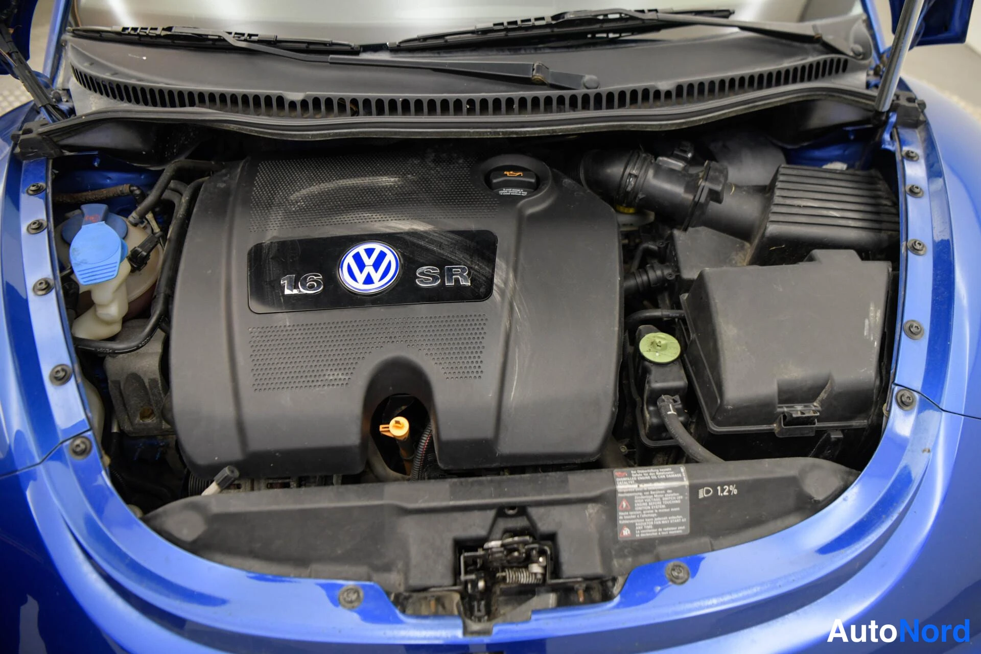 Muu: Moottoritilan yläkuvassa Volkswagenin moottorisuojus, jossa on teksti 1.6, VW-tunnus ja SR; sinisen korirakenteen reunat näkyvät laidoissa.