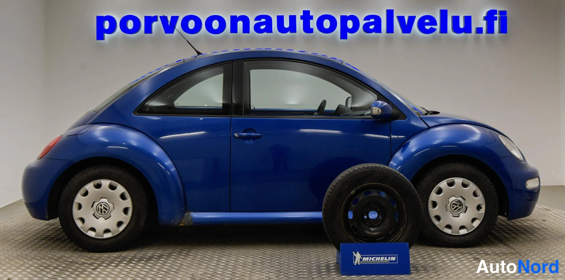 Etuviisto, nokka oikealle: Sininen Volkswagen New Beetle kuvattu sivulta hieman keulakulmasta, kylki ja etuvalo näkyvät; renkaita edessä lattialla.