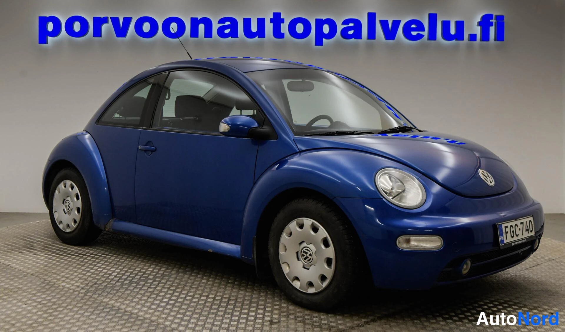 Etuviisto, nokka oikealle: Sininen Volkswagen New Beetle kolmannelta kulmalta keula oikealla, kylki ja etuosa näkyvät, rekisterikilpi.