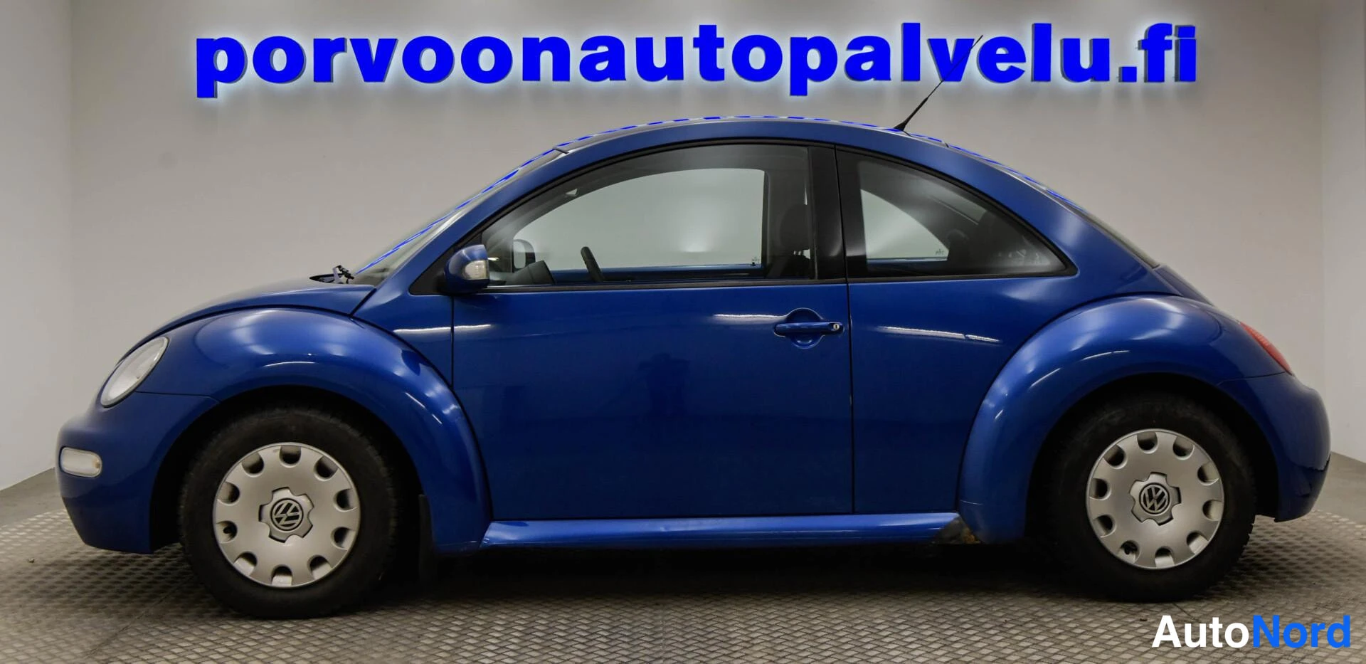 Etuviisto, nokka vasemmalle: Sininen Volkswagen New Beetle kolmannelta kulmalta keula vasemmalla, kylki ja etuvalo näkyvät.