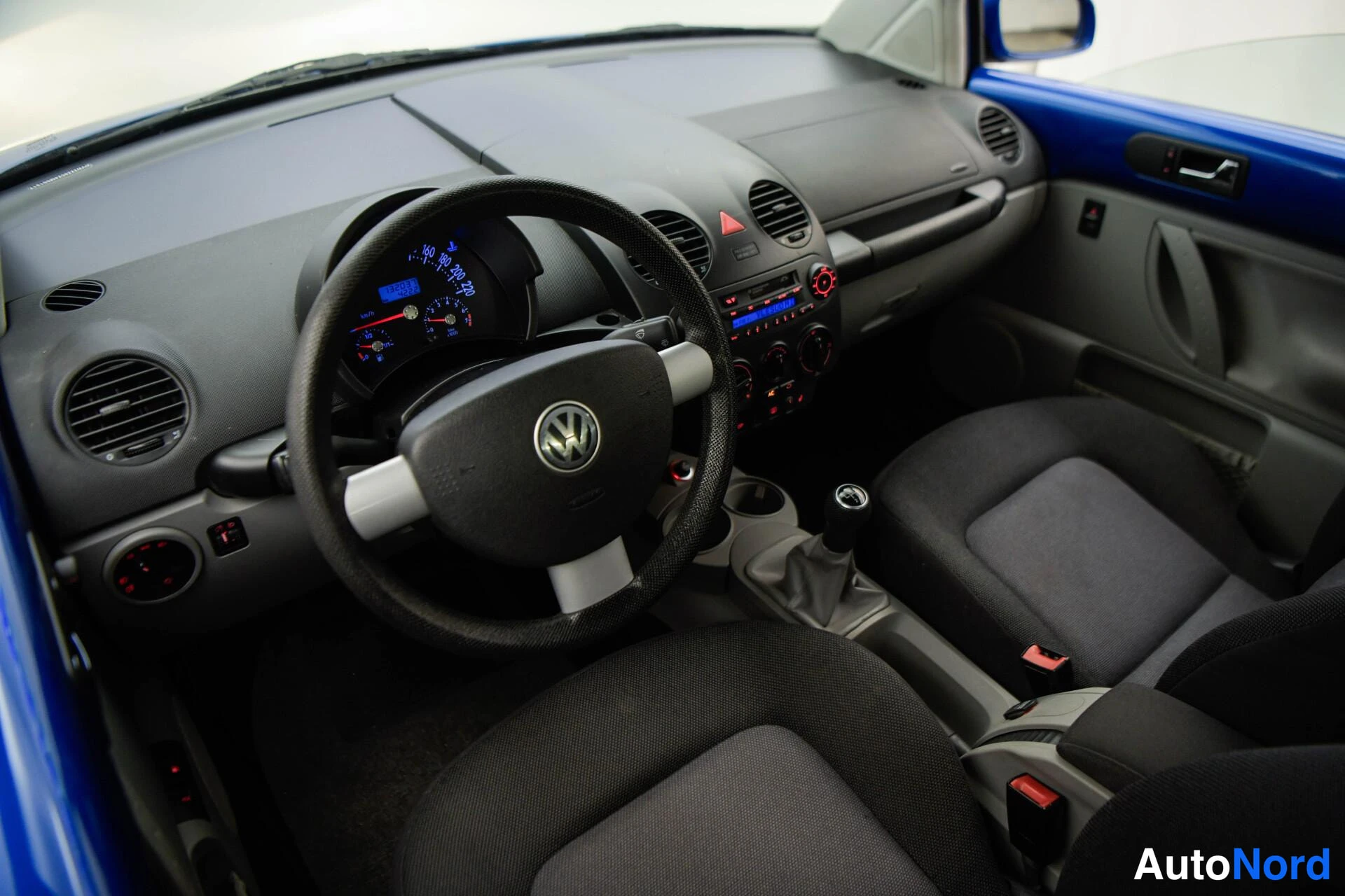 Edestä: Auton sisätilat: pimeä kojelauta, VW-ohjauspyörä, manuaalivaihtopyörä ja kaksi tummaa istuinta.