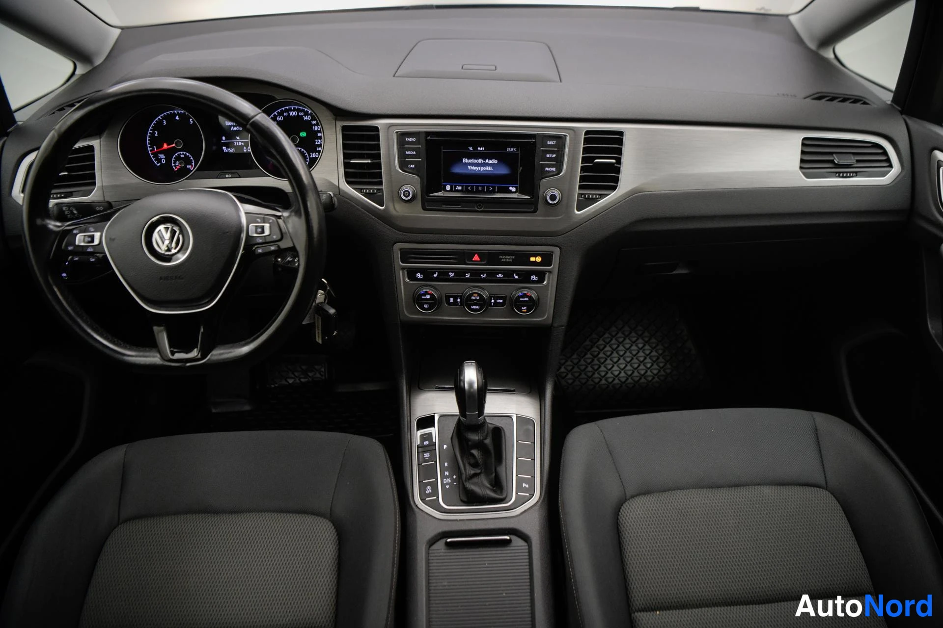 Yksityiskohta: Ohjaamo näkyy edestä: VW-merkkinen monitoimiohjaus, mittaristo, kosketusnäytöllinen radio, automaattivaihteen valitsin ja kaksi etuistuinta.