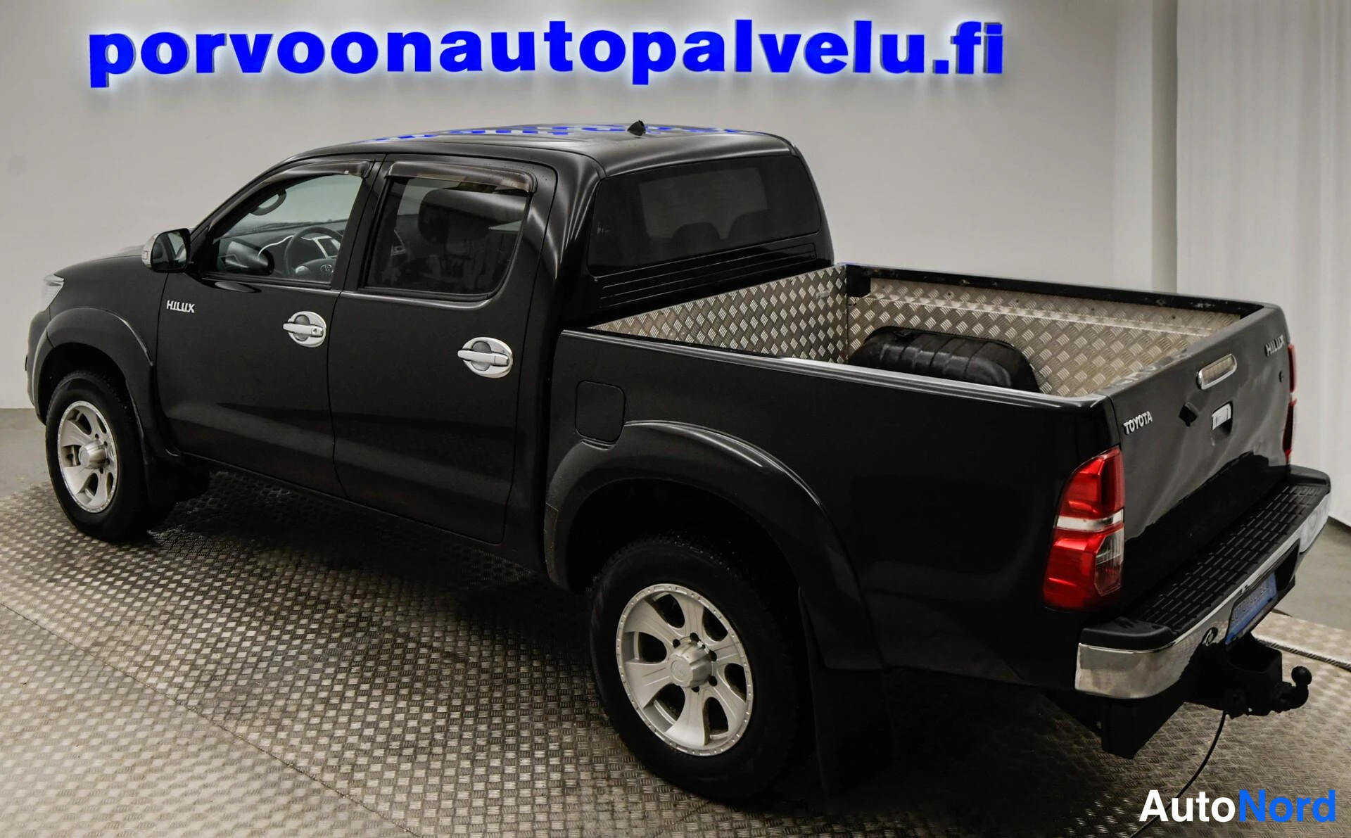 Takaviisto: Mustan Toyota Hilux -lainen pakettiauto takaa vasemmalta kulmasta kuvattuna, takavalo ja vasen kylki näkyvissä.