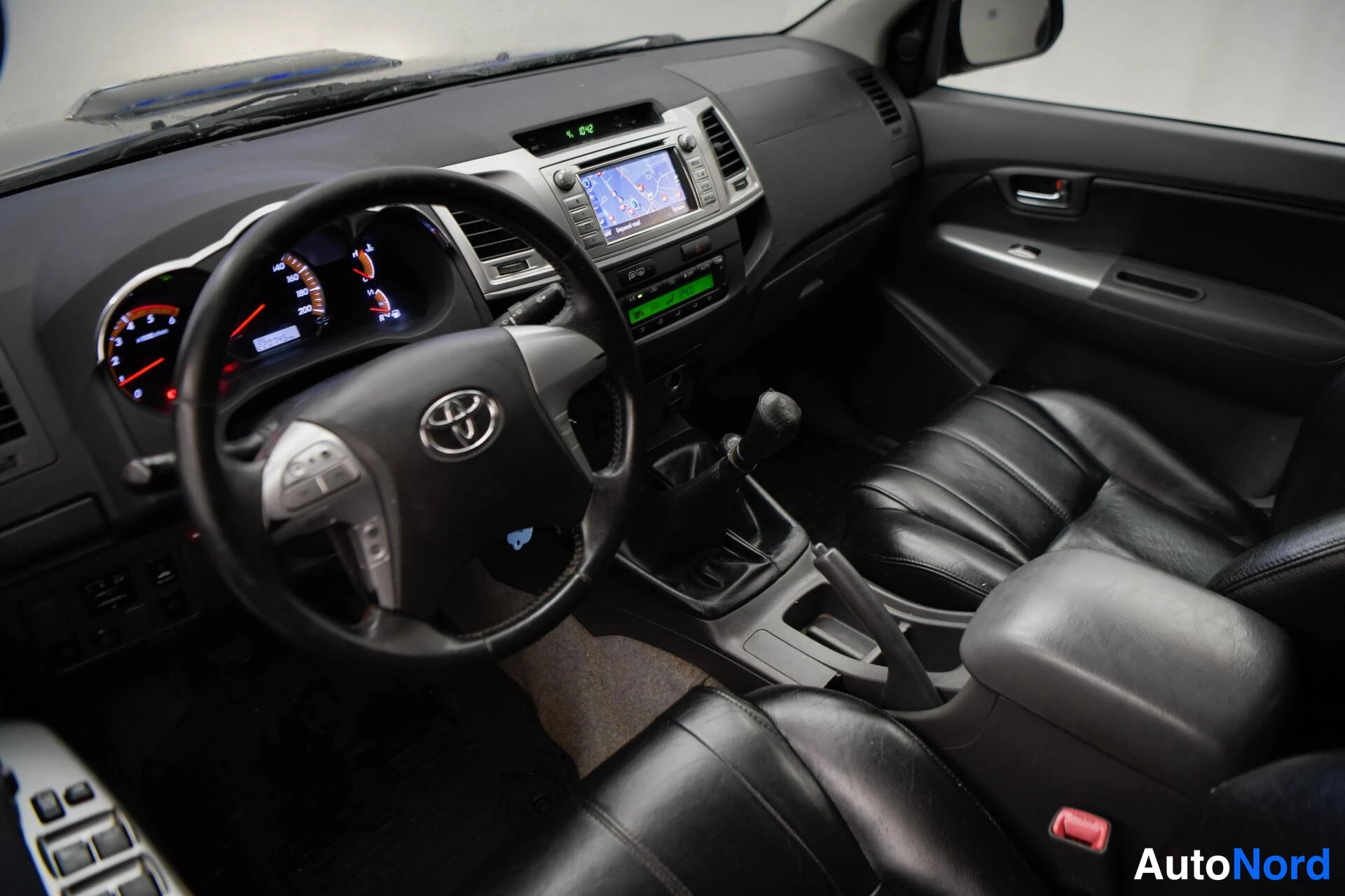 Edestä: Sisäkuva etuistuimilta: Toyota-ohjauspyörä, mittarit, karttanäyttö kojelaudassa, manuaalivaihto ja mustat nahkaistuimet.