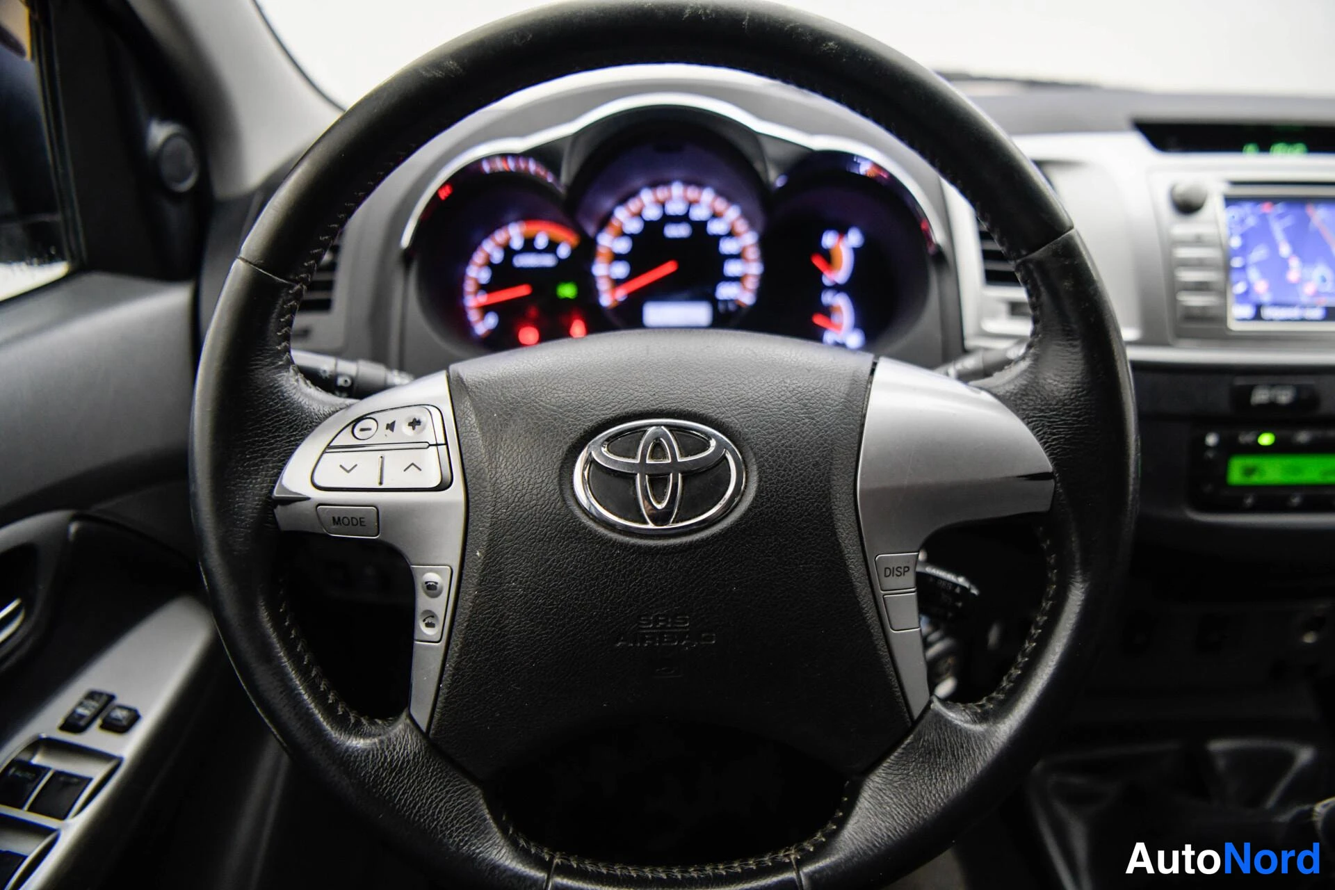 Edestä: Lähikuva Toyota-ohjauspyörästä ja mittaristosta; ohjauspyörän napissa SRS Airbag -merkintä ja ääniohjaimet vasemmalla puolella.