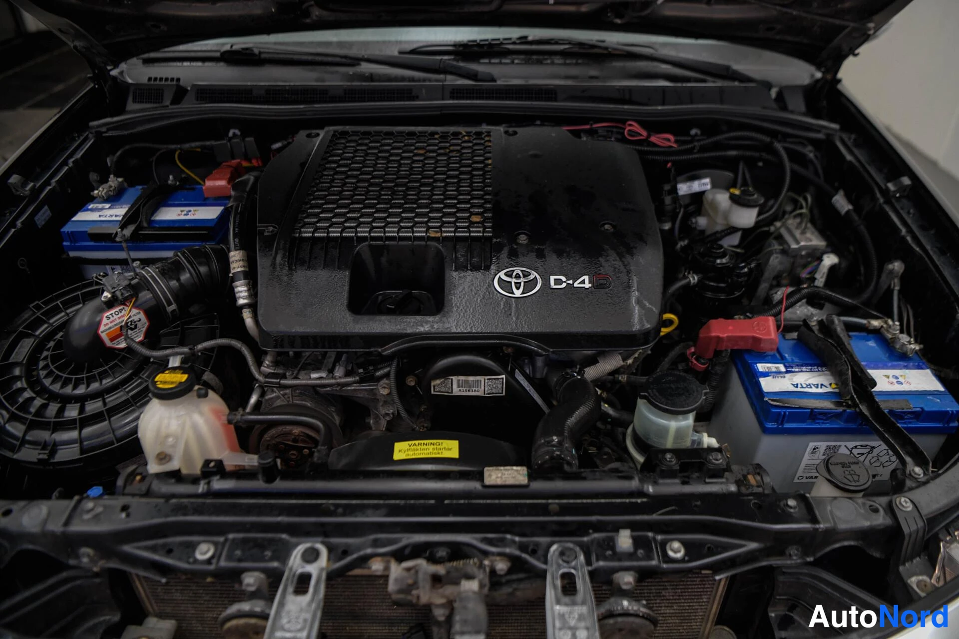 Muu: Moottoritila kansi auki; moottorin suojakannessa Toyota-logo ja D-4D-teksti, Varta-merkkinen akku näkyvissä.