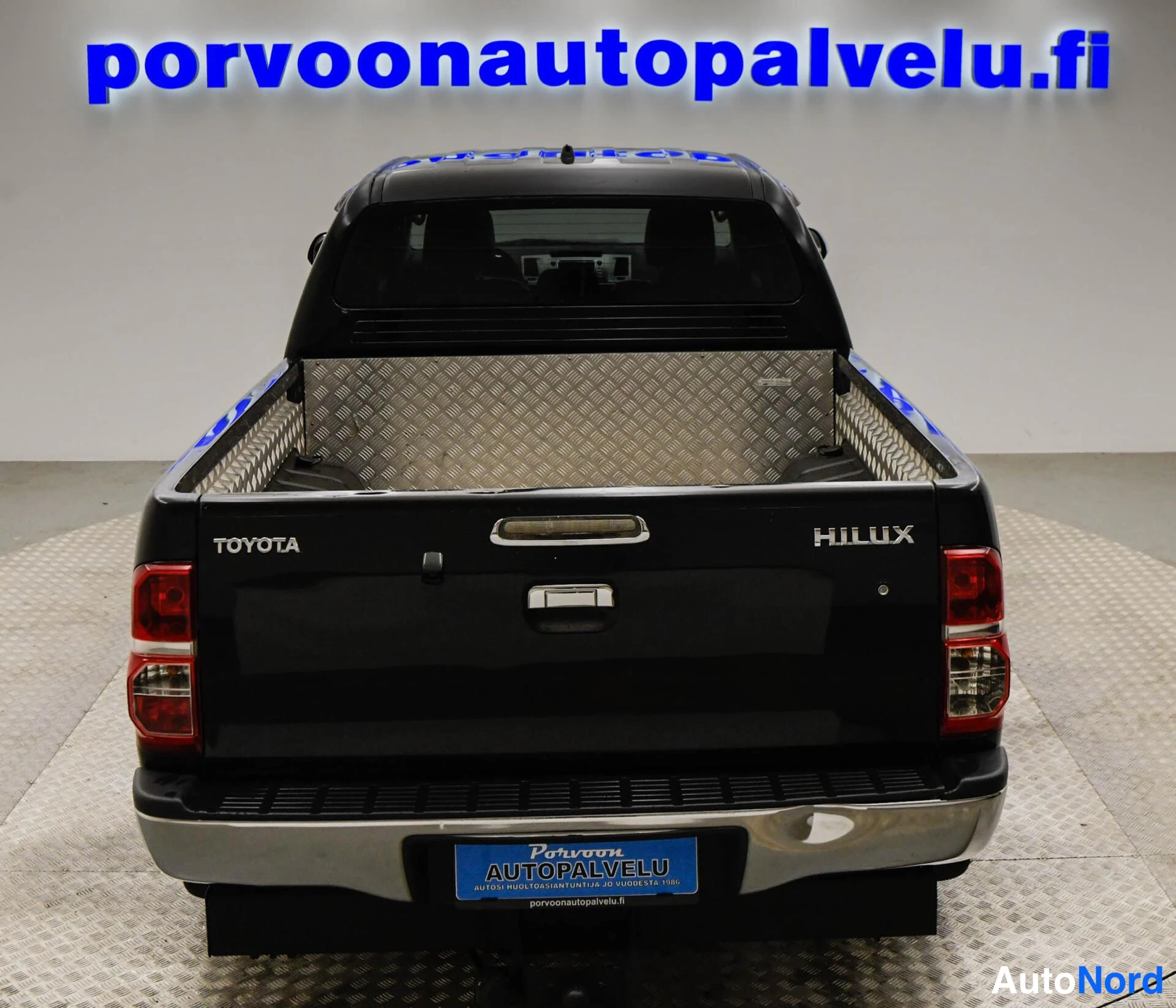 Takaa: Mustan Toyota Hilux -pakettiauton takaosa suoraan takaa, TOYOTA- ja HILUX-tekstit sekä takaosalasi näkyvät.