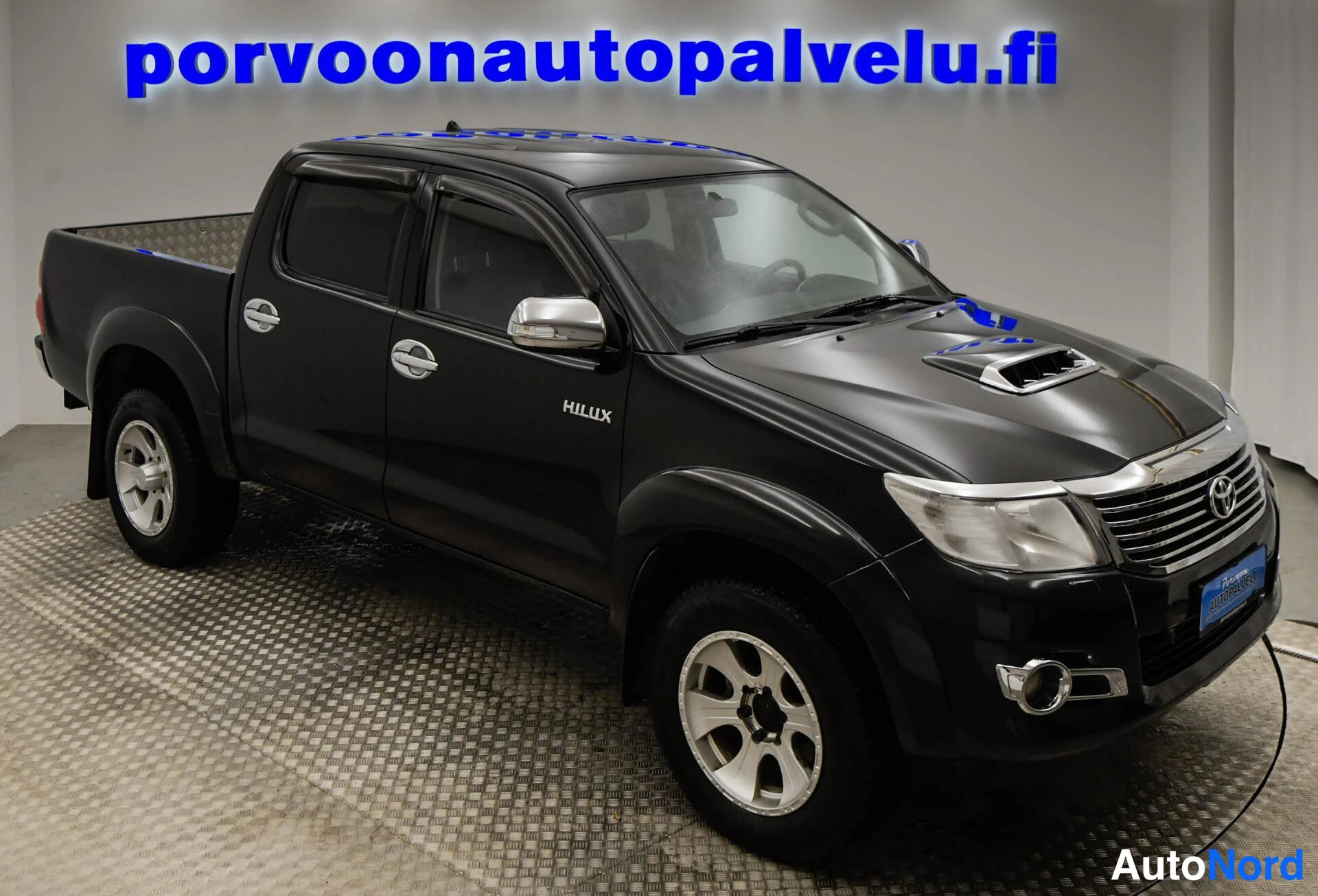 Etuviisto, nokka oikealle: Musta Toyota Hilux etuviistosta oikealta, kylki ja etuposetti näkyvissä autonäyttämöllä.