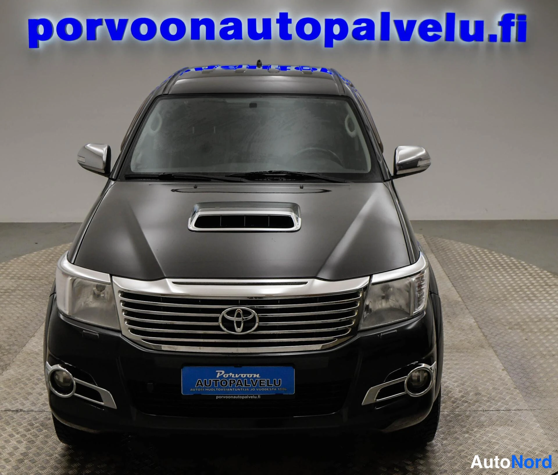 Edestä: Mustan Toyota Hiluxin etunäkymä suoraan edestä, kromiset ilmastointiaukot ja Toyota-merkki näkyvissä.