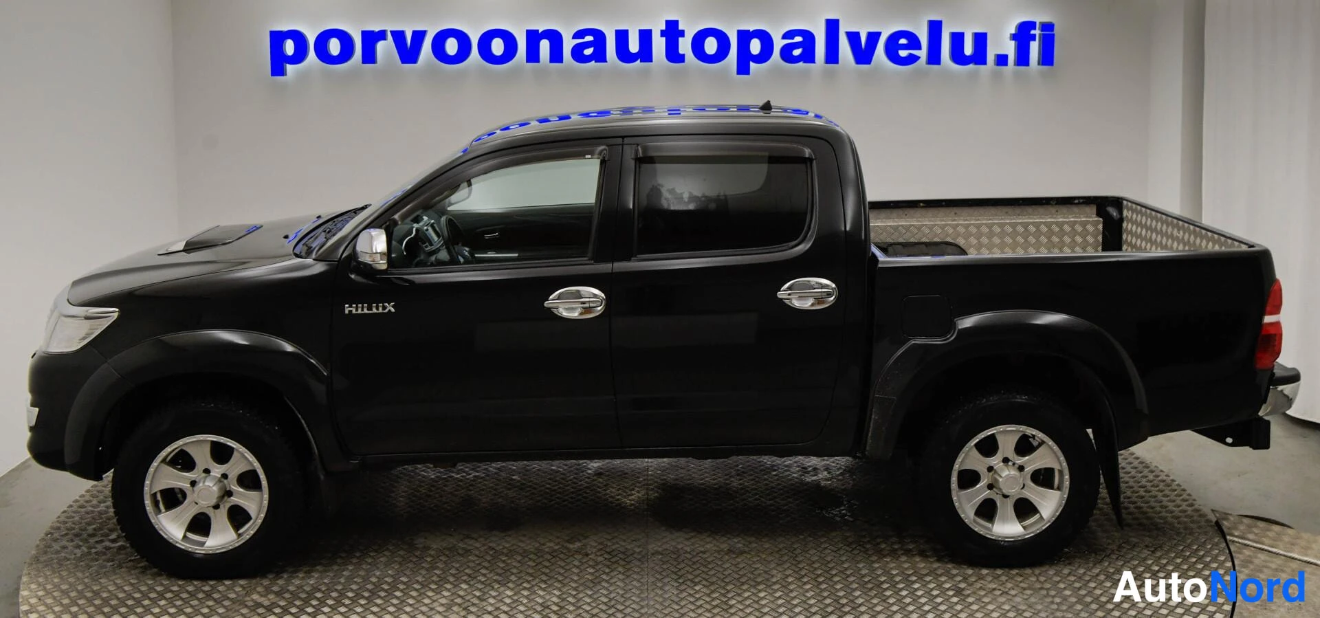 Sivulta: Musta Toyota Hilux -avolava sivuprofiilissa oikealta katsottuna sisätiloissa.