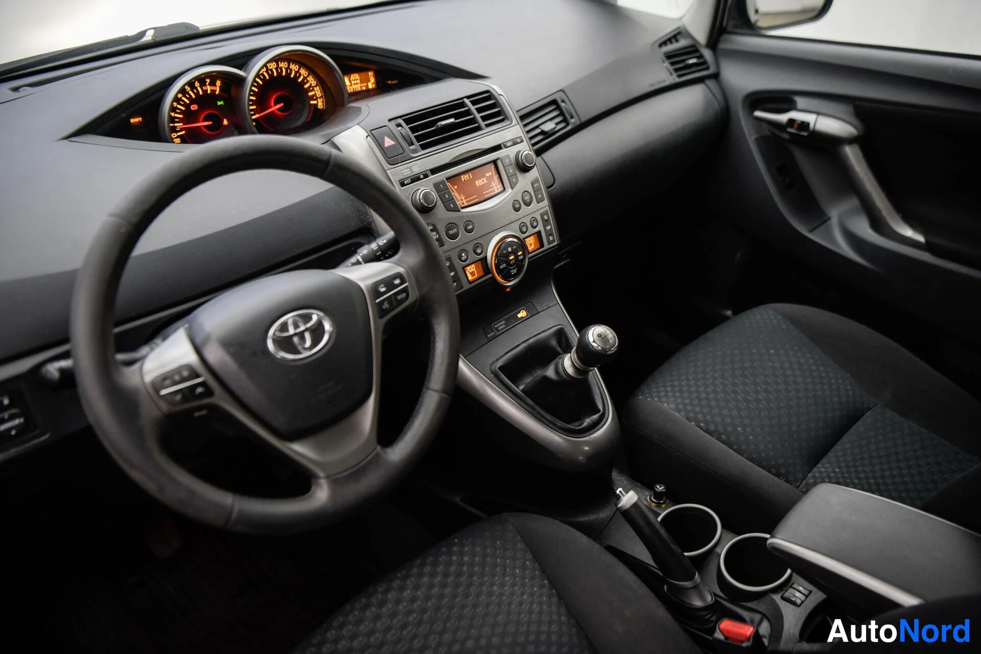 Sivulta: Ohjaamon yleiskuva kuljettajan puolelta: Toyota-ohjauspyörä, mittaristo, radio ja manuaalivaihto keilalla.