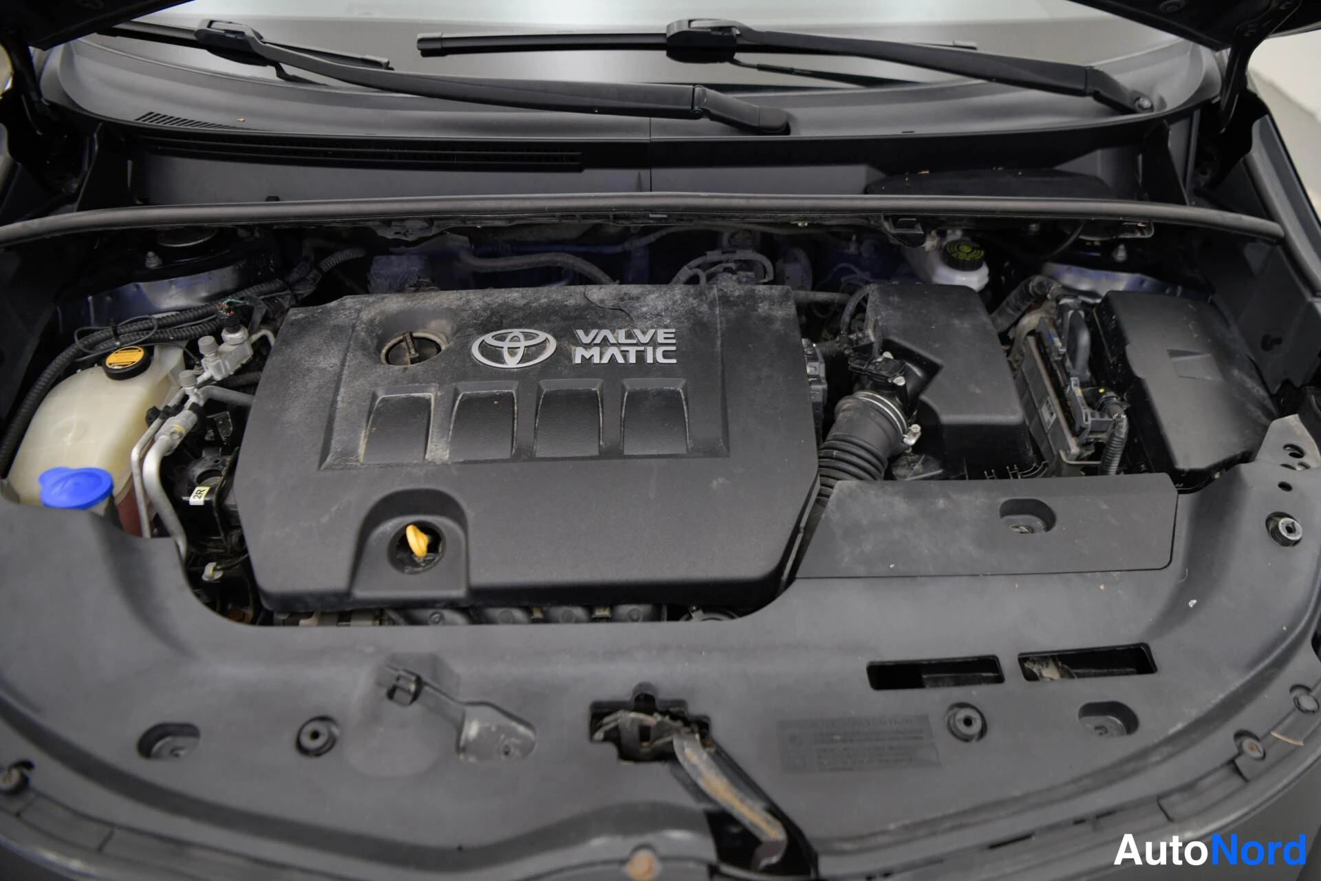 Muu: Avaamaton moottoritila: musta konepelti ja moottorin suojus Toyota-logo ja tekstillä VALVE MATIC, jäähdytinneste- ja pesunestesäiliöt näkyvissä.