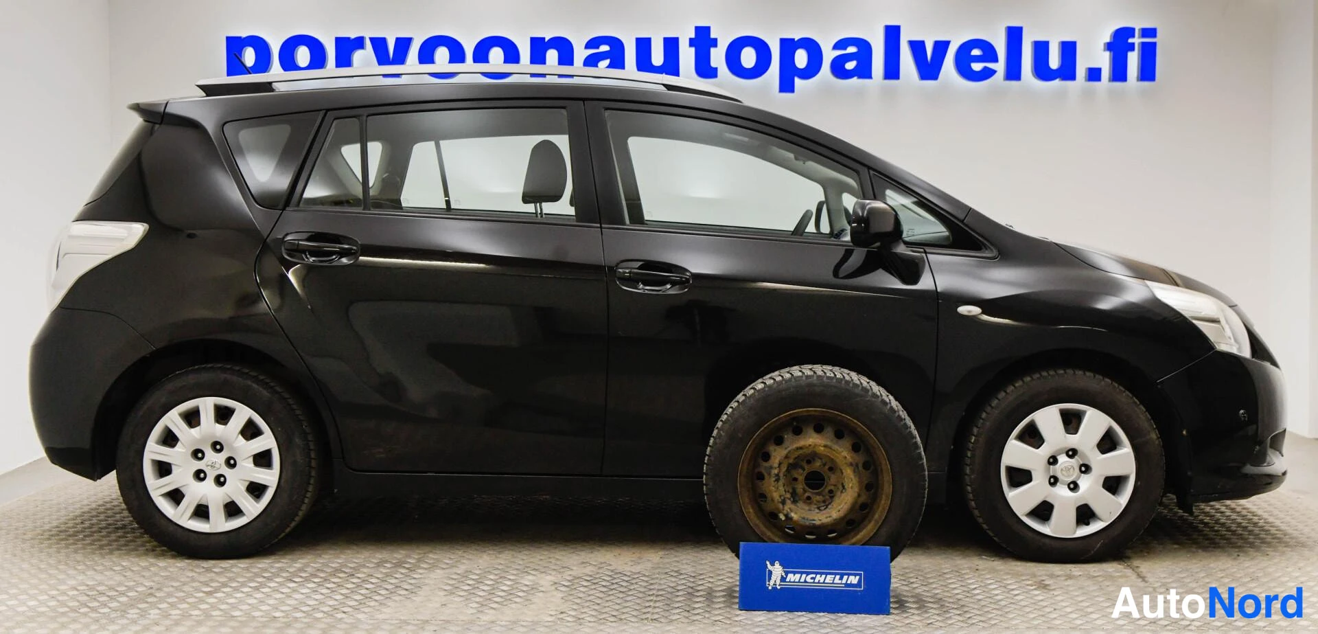 Sivulta: Mustan tila-auton sivuprofiili autotallissa, sininen porvoonautopalvelu.fi-kyltti taustalla ja irtonainen rengas ja vanteenkehys lattialla auton edessä.