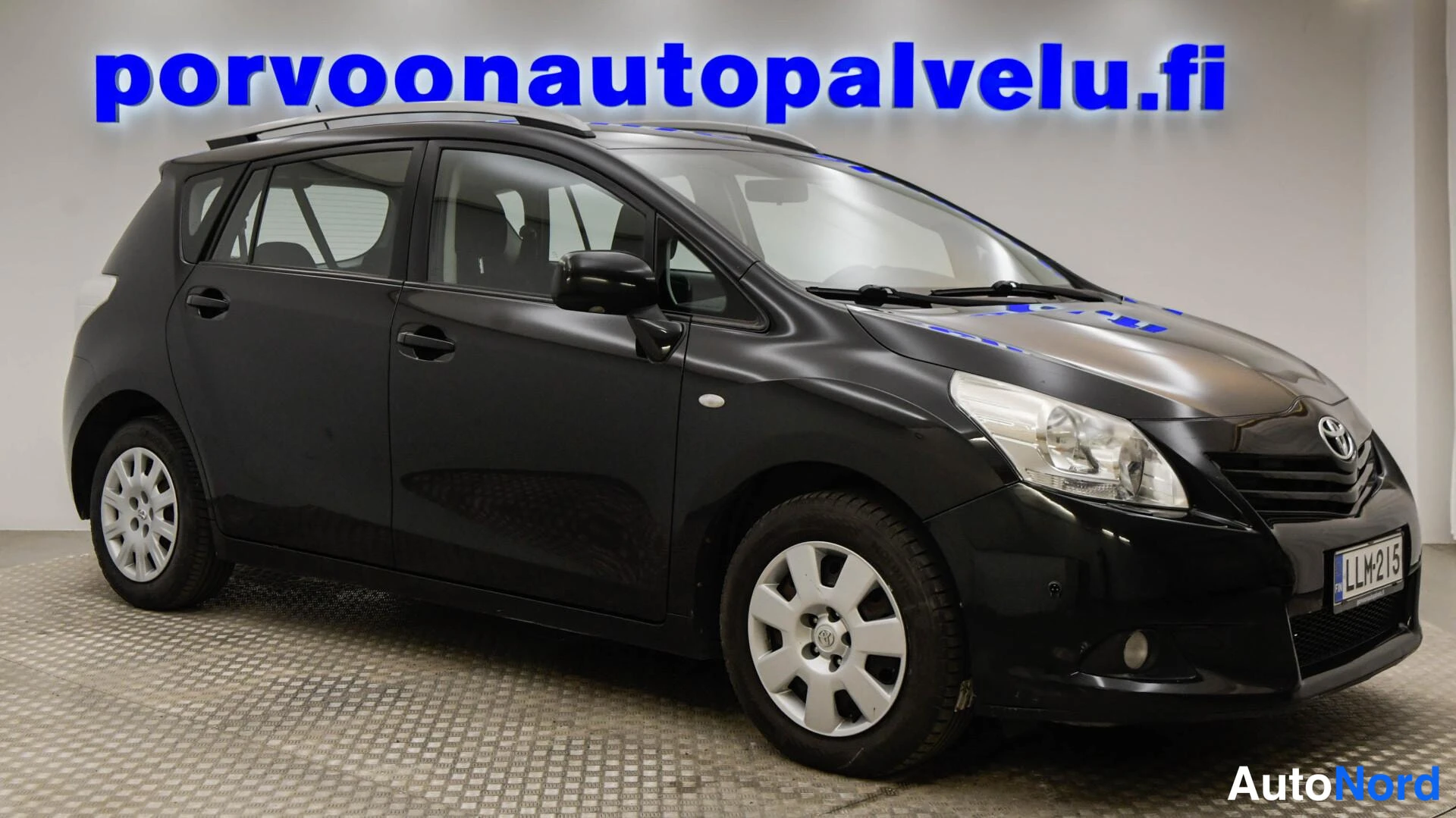 Etuviisto, nokka oikealle: Mustan tila-auton etu-kolmasosakuva oikealta, Toyota-merkki konepellin etupyörien vanteet näkyvät, taustalla sininen porvoonautopalvelu.fi-kyltti.
