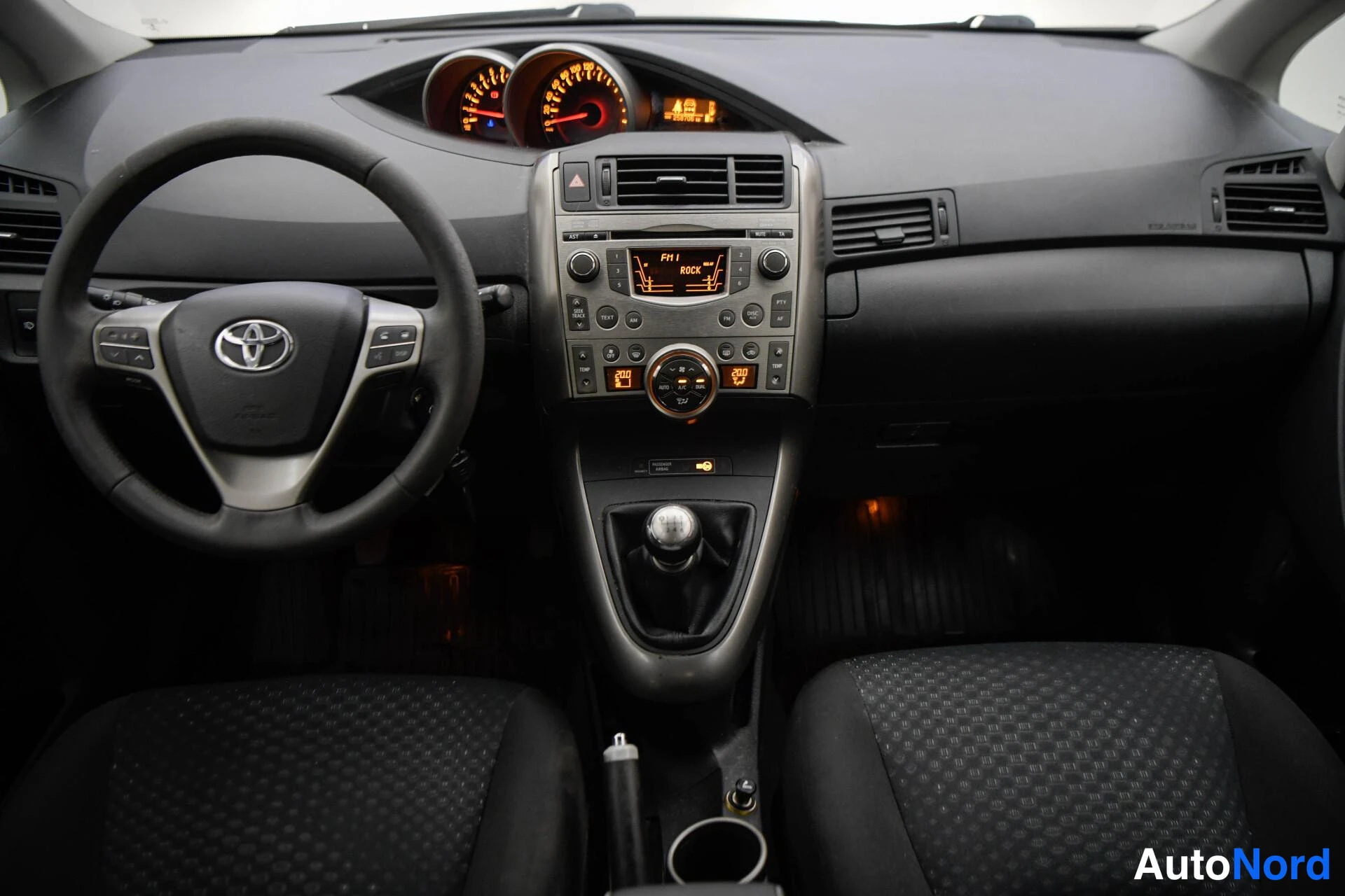 Sivulta: Kohtisuora kuva mittaristosta ja keskikonsolista: Toyota-ohjauspyörä, radiopaneeli näytöllä ja manuaalivaihto keilalla.