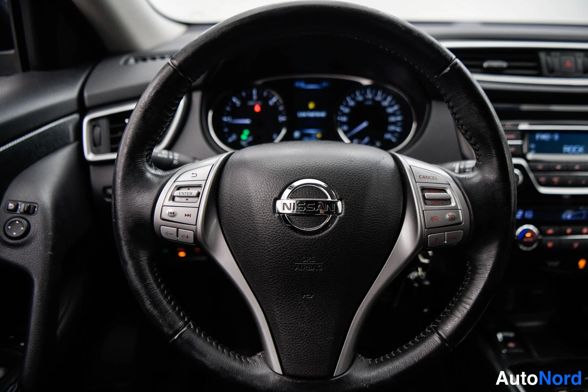 Muu: Nissan-ohjauspyörän lähikuva: keskiairbag-merkintä ja monitoiminapit, mittaristo ja radiopaneeli taustalla.