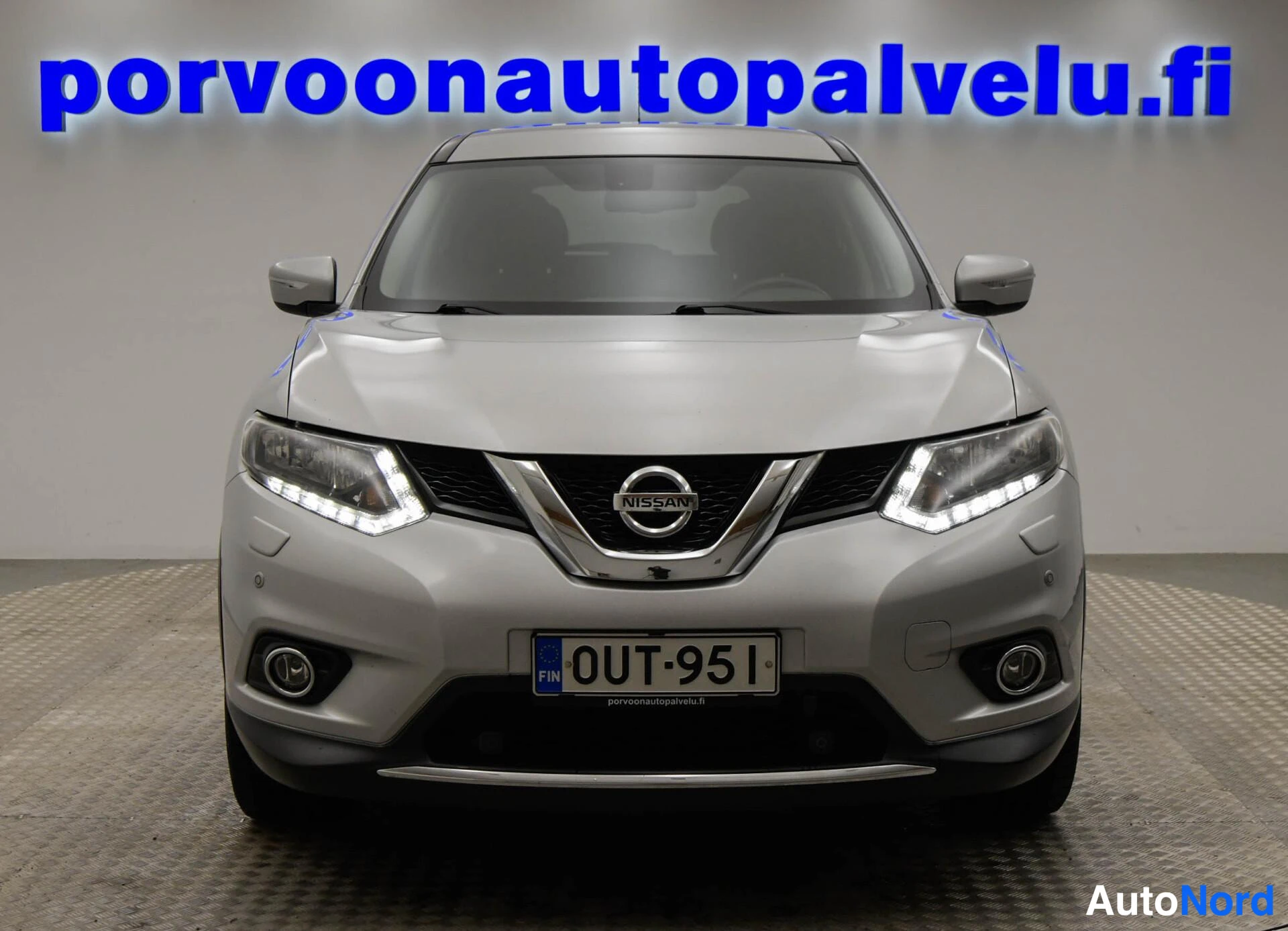 Edestä: Auto suoraan edestäpäin: hopearunkoinen, X-trail-merkki konepellissä, rekisterikilpi.