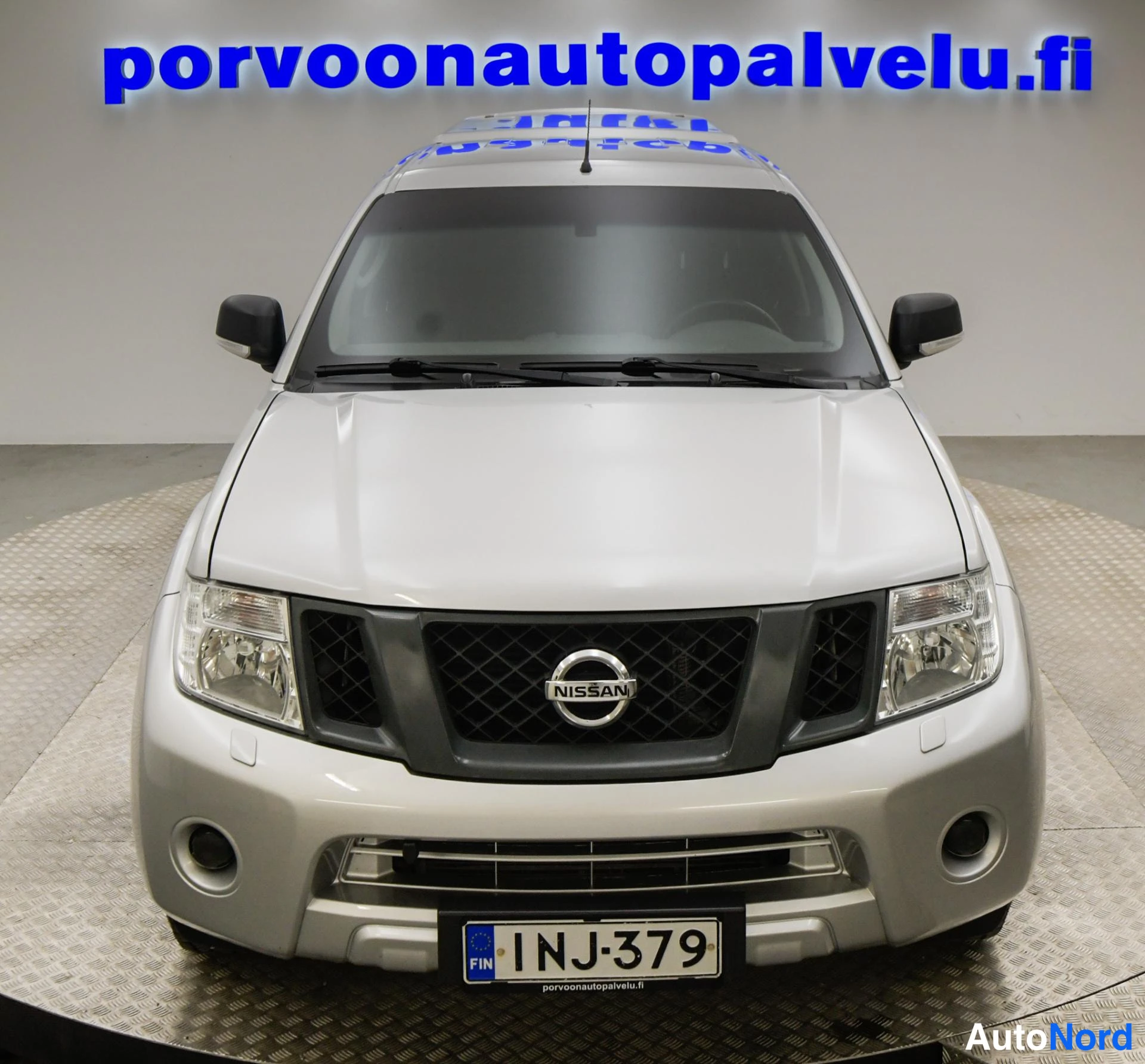 Edestä: Hopeanvärinen nelistäauto suoraan edestä kuvattuna; säleikössä Nissan-merkki ja rekisterikilpi näkyvät.