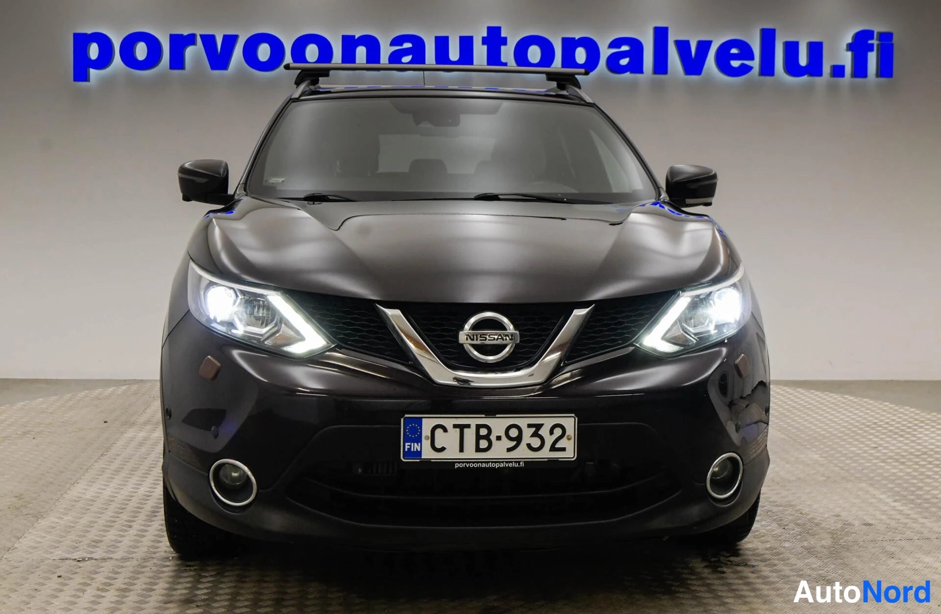 Edestä: Suora etunäkymä; tumma Nissan Qashqai, kattotelineet, rekisteri, etutuleissa takuvalot päällä.
