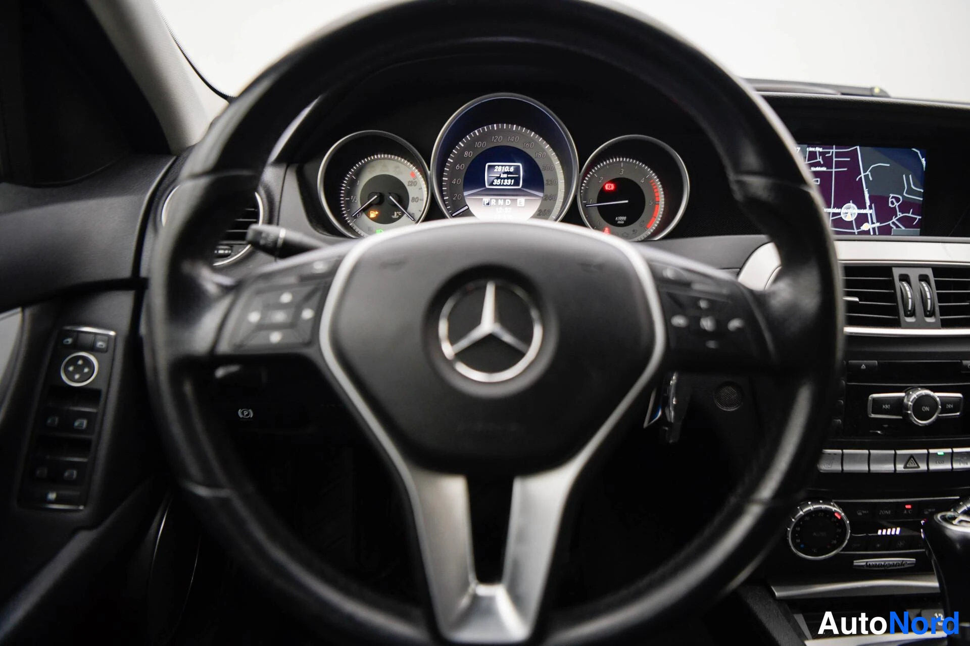 Muu: Lähikuva Mercedes-Benzin ohjauspyörästä ja mittaristosta; nopeusmittari ja kierroslukumittari erottuvat, karttanäyttö osittain näkyvissä oikealla.