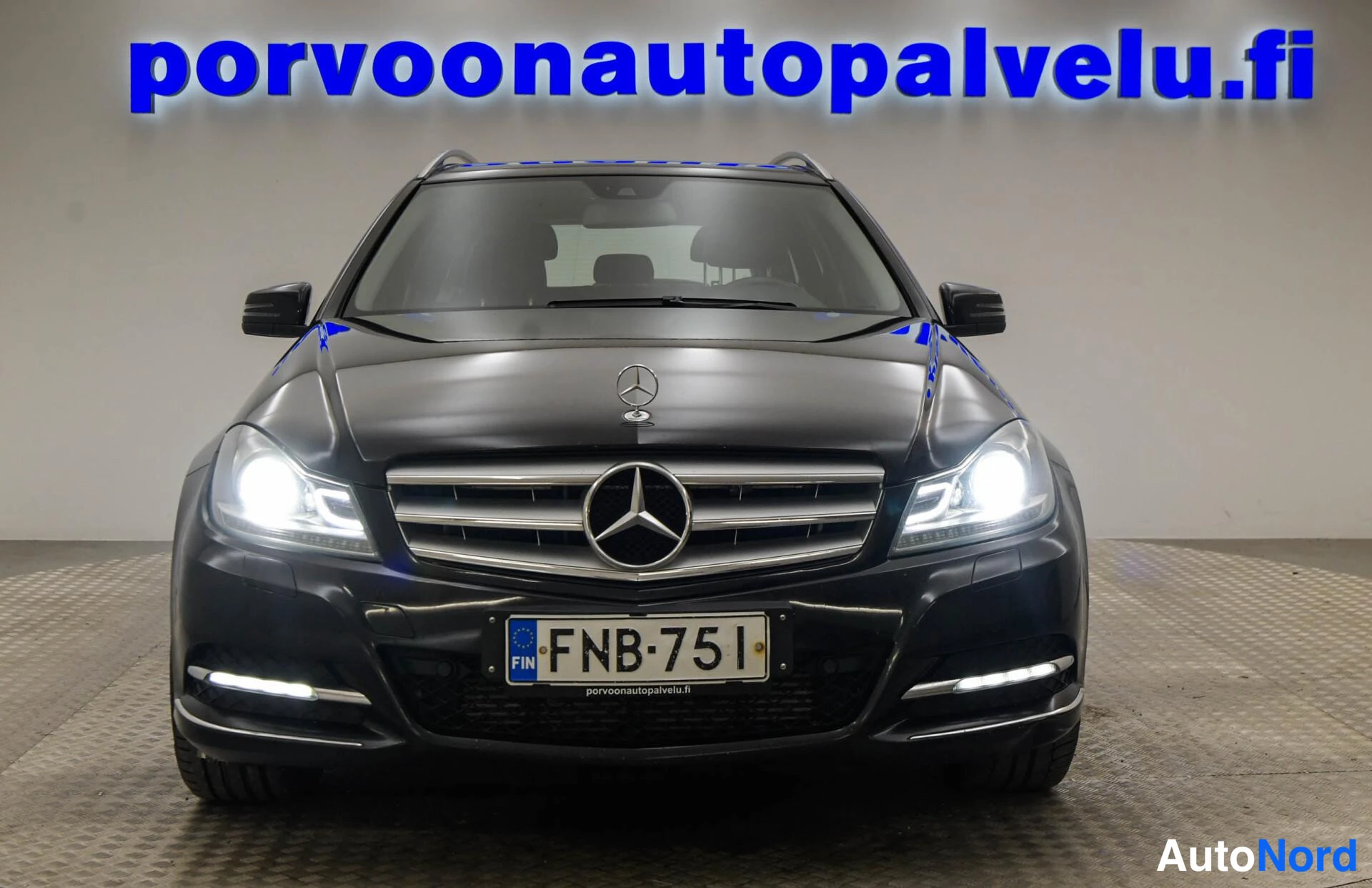 Edestä: Mustan Mercedes-Benz C-sarjan farmarin suora etunäkymä, galvanoidussa kyltillä rekisterinumero, ajovalot päällä.