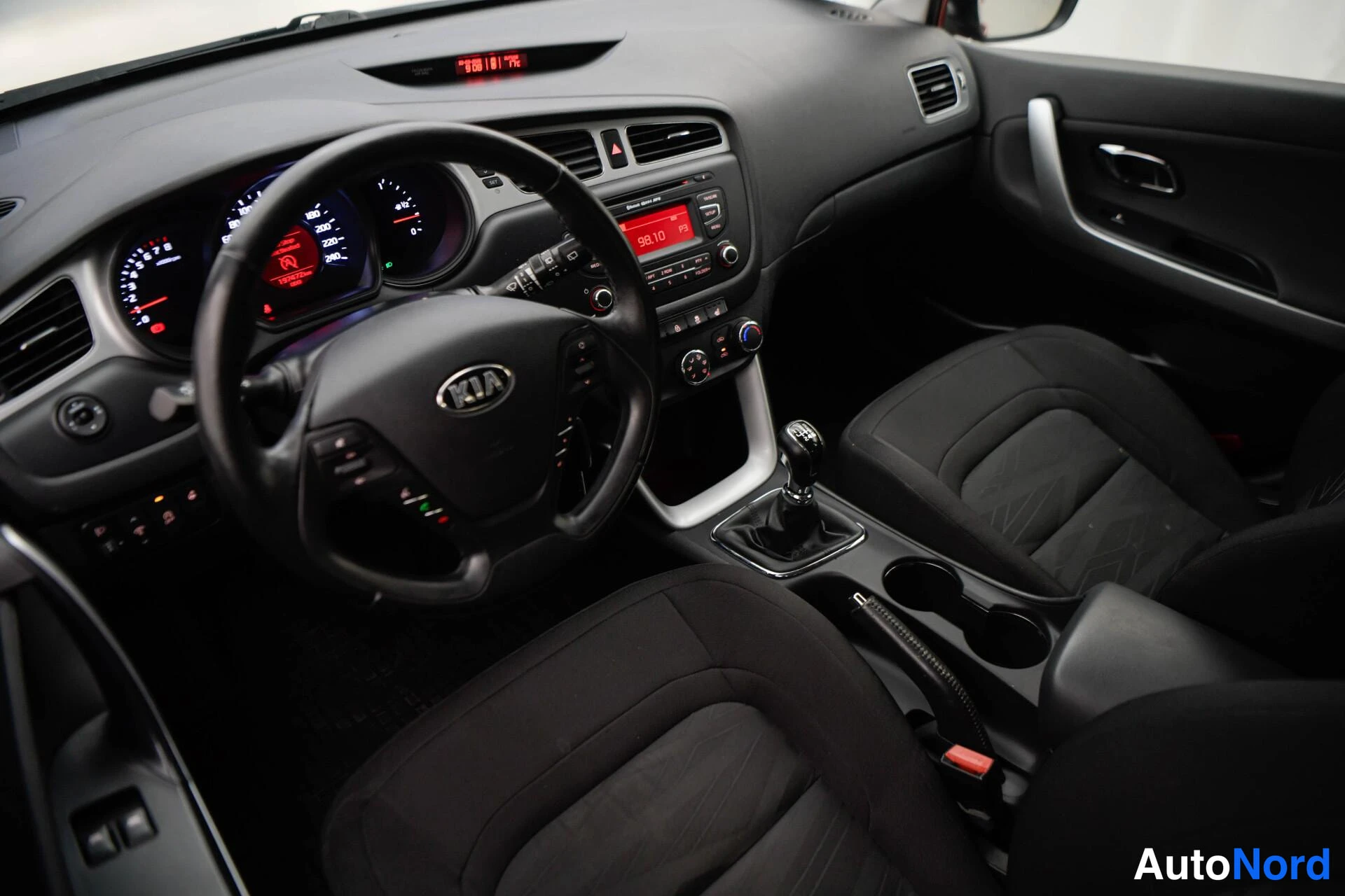 Edestä: Kia Ceedin ohjaamo ylhäältä: KIA-ohjauspyörä, mittaristo, radio näytöllä 98.10, manuaalivaihto ja tummat penkit.