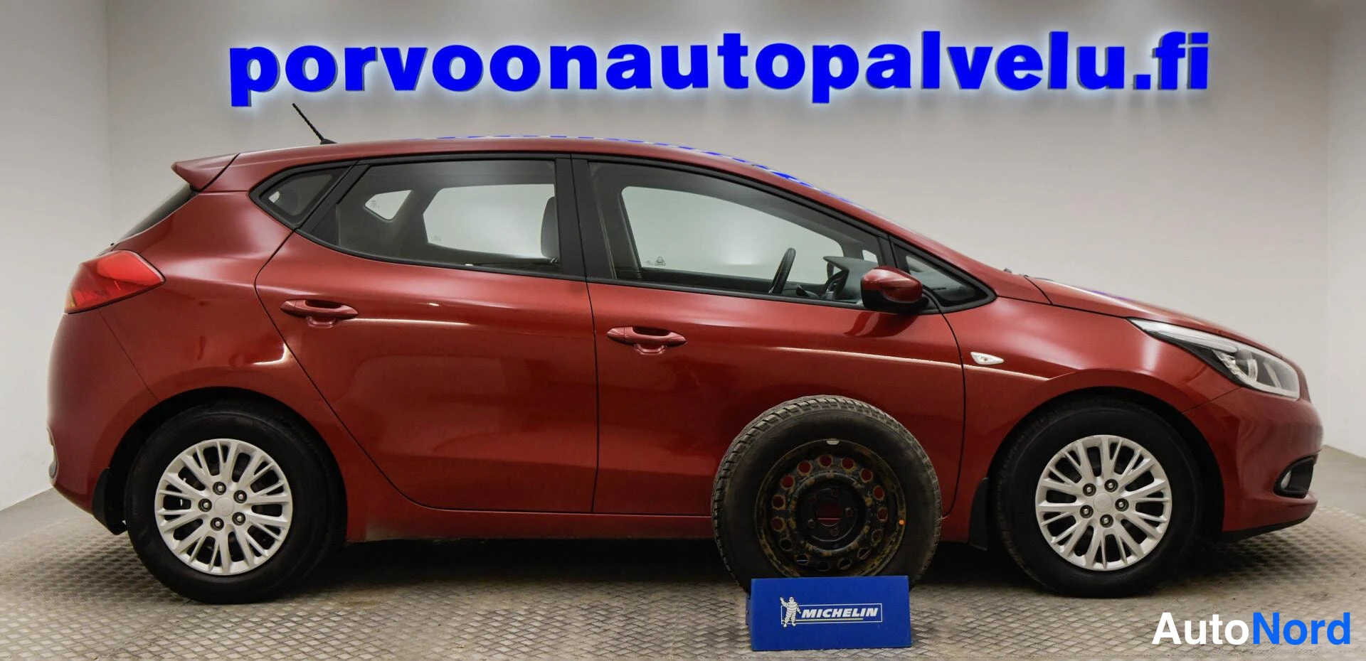 Etuviisto, nokka oikealle: Punainen Kia Ceed sivukulmassa sisätiloissa, auton etupuolella lattialla varapyörä ja sininen Michelin-kyltti.