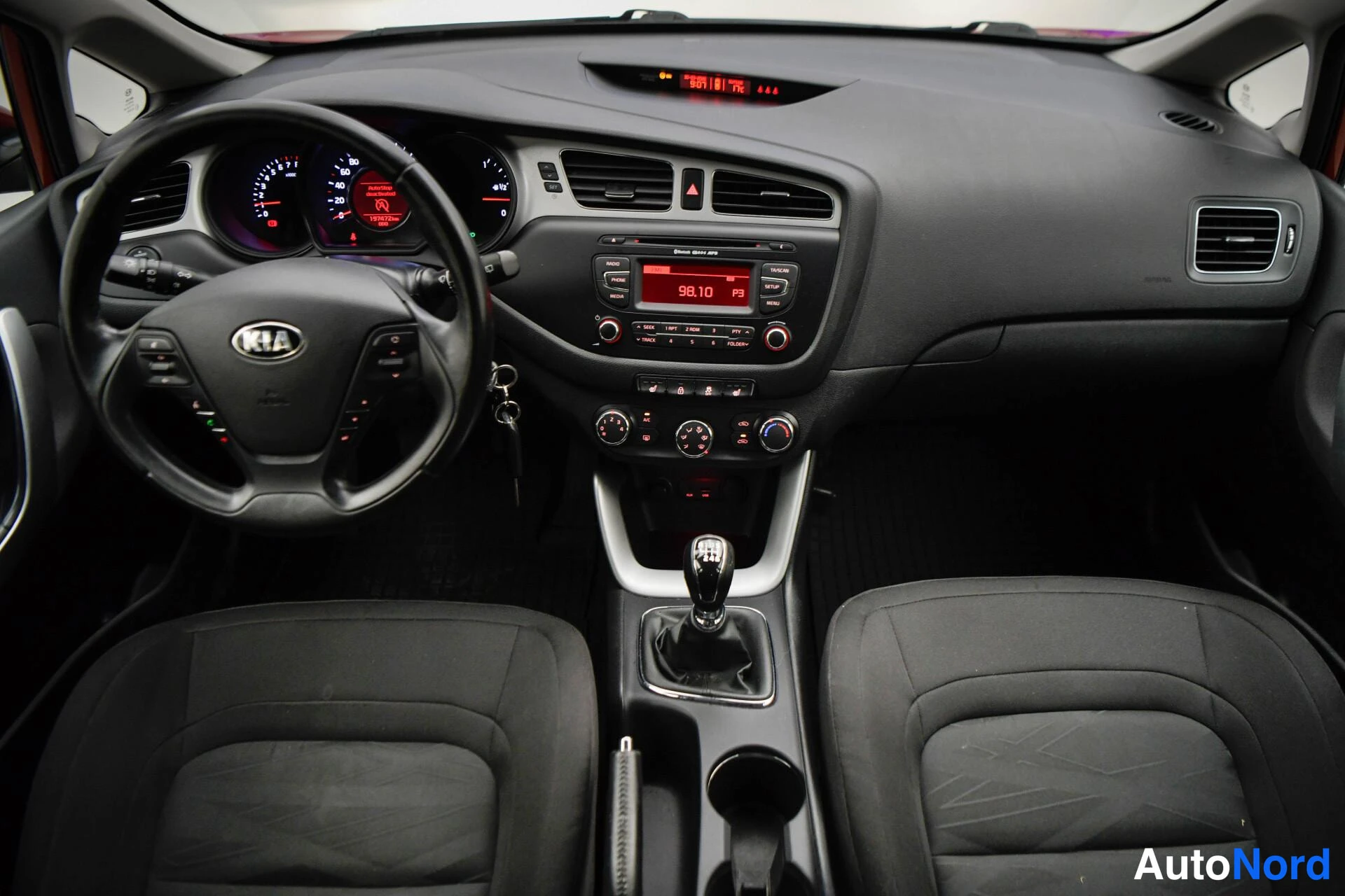 Edestä: Kia Ceedin ohjaamo: KIA-merkitty ohjauspyörä, analogiset mittarit, radio näytöllä 98.10, manuaalivaihtehallitsija ja käsijarru.