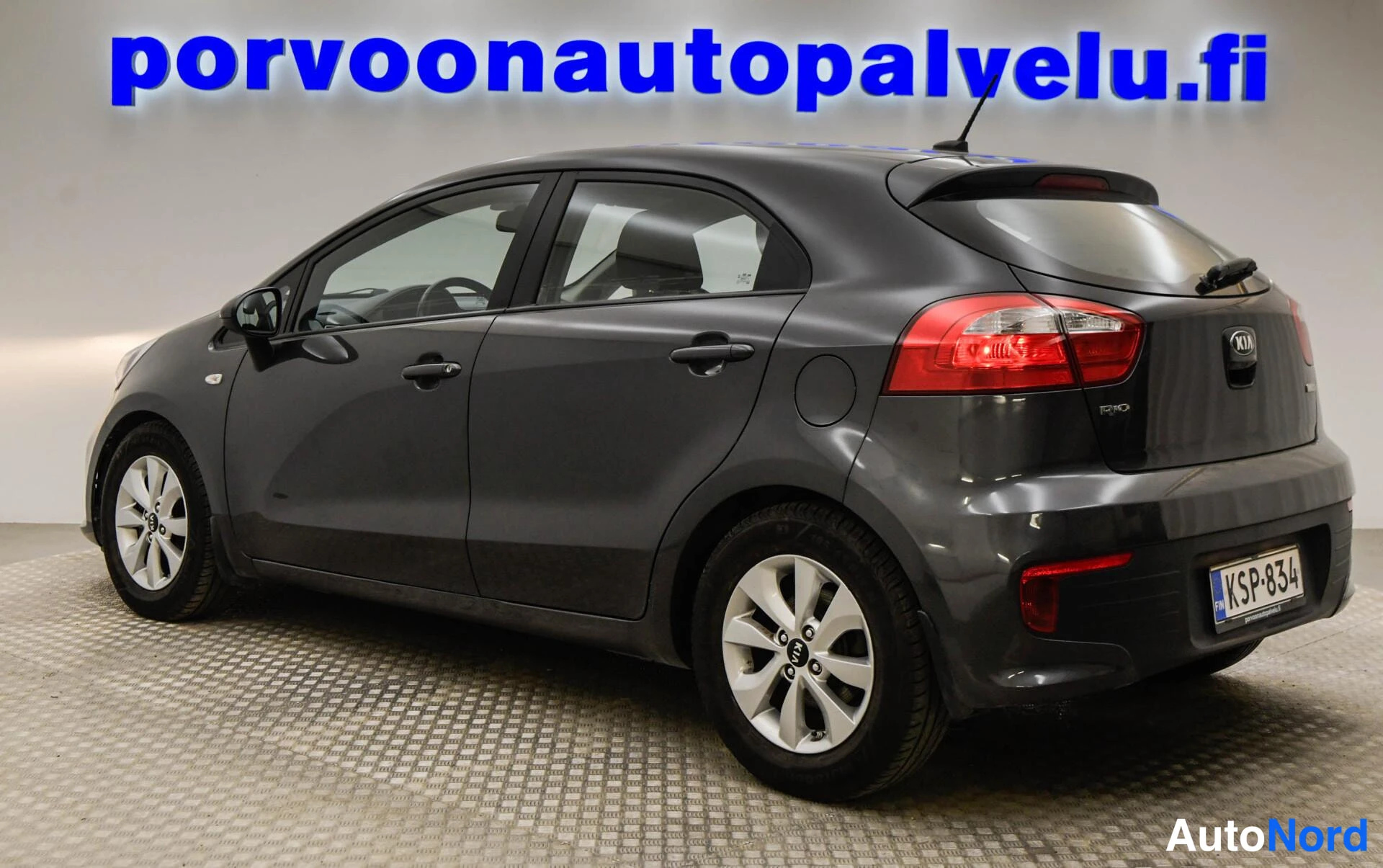Takaviisto: Kia Rio -viistokompakti takakulmasta oikealle, tummanharmaa kori, -rekisterikilpi ja porvoonautopalvelu.fi-taustalogo.