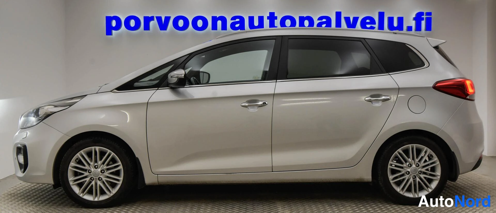 Takaviisto: Hopeanvärinen Kia Carens takaovet ja takavalo näkyen, sivuseinä autoa vasten.
