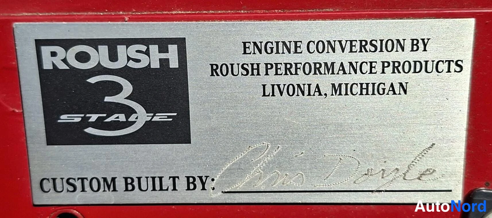 Yksityiskohta: Metallikilpi punaisella pohjalla: ROUSH 3 STAGE -logo sekä teksti 'ENGINE CONVERSION BY ROUSH PERFORMANCE PRODUCTS LIVONIA, MICHIGAN' ja 'CUSTOM BUILT BY' allek