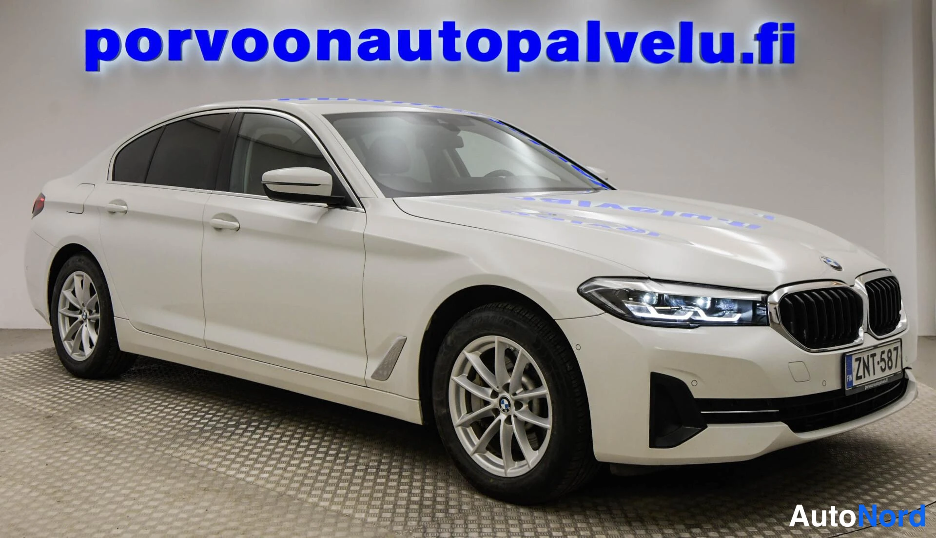 Etuviisto, nokka oikealle: Valkoinen BMW-sedan kolmineljänneskuva keula oikealla; rekisterikilpi.