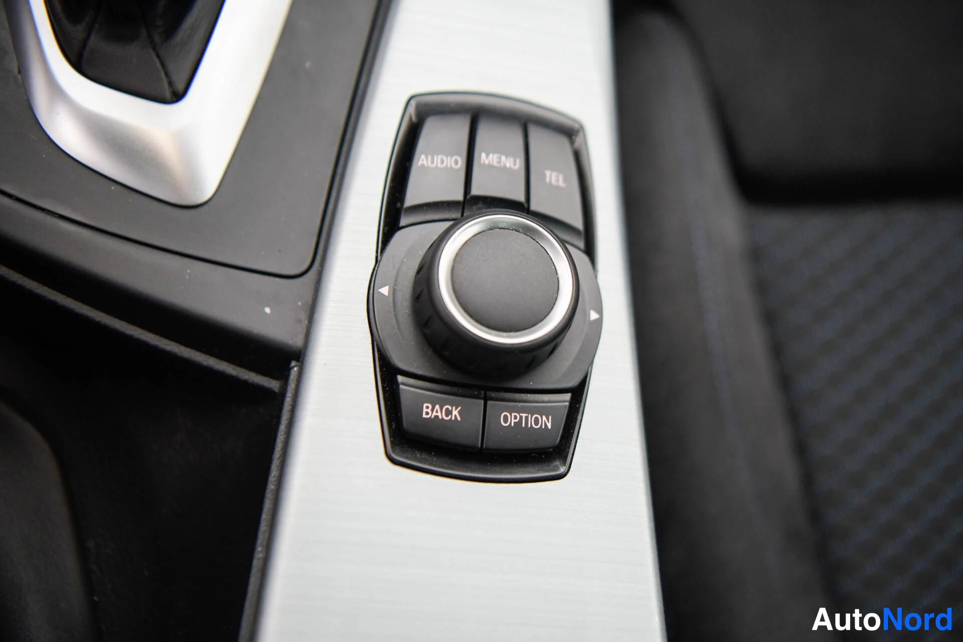 Etuviisto, nokka vasemmalle: Lähikuva iDrive-ohjaimesta: pyöreä ohjainnuppi ja painikkeet Audio, Menu, Tel, Back ja Option alumiinikoristeisella panelilla.