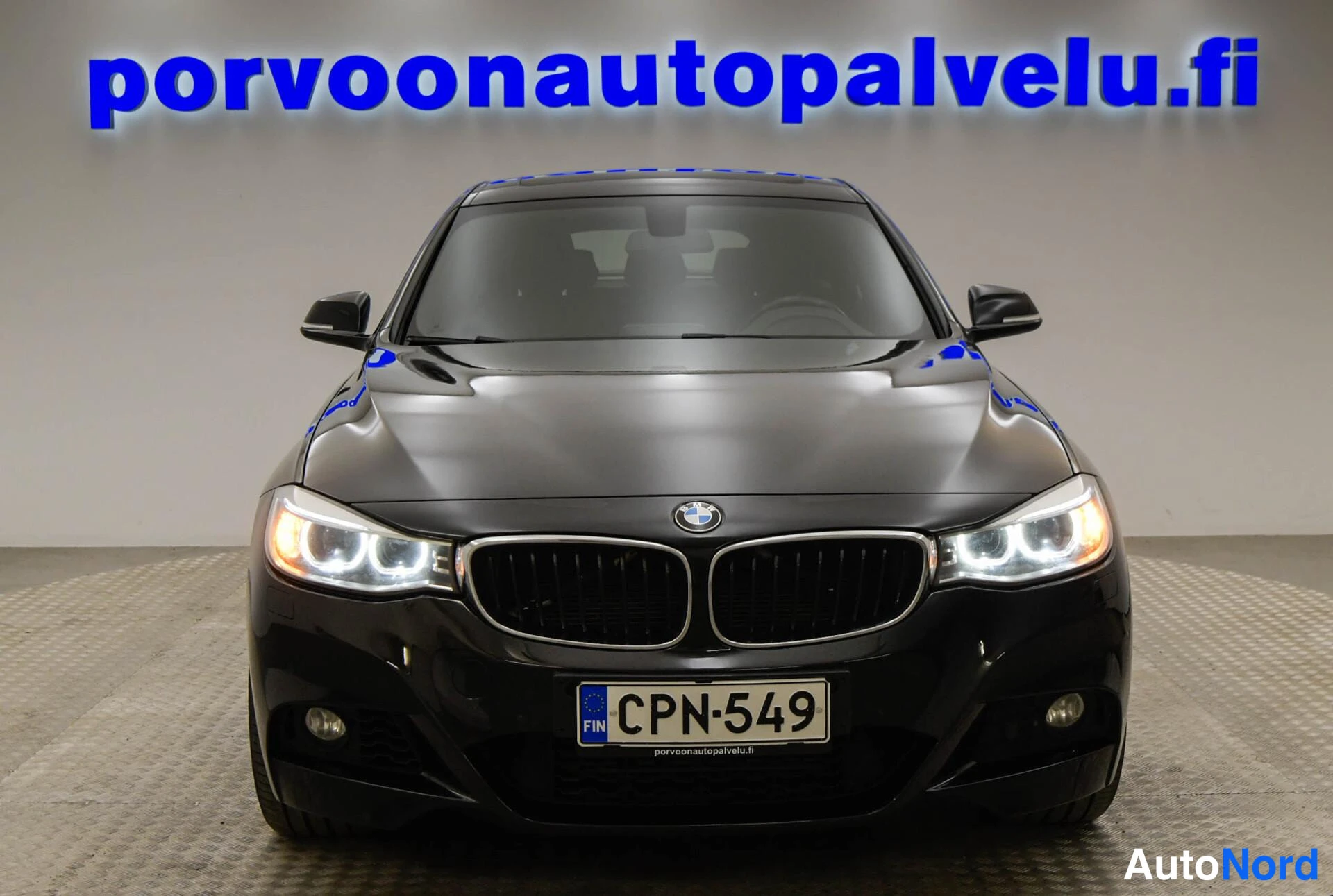 Edestä: Mustan auton etunäkymä, etuvilkut palaa, rekisteritunnus ja BMW-merkki säleikössä.
