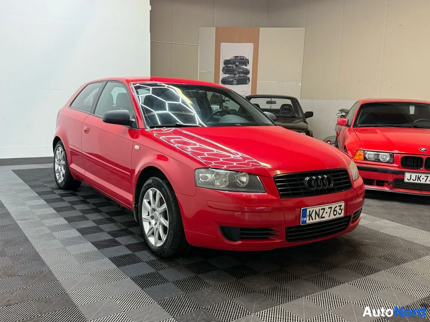 Etuviisto, nokka oikealle: Punainen Audi A3 -viistoperä kolmesta neljänneksestä edestä oikealta, takana toinen punainen henkilöauto, sisätiloissa.