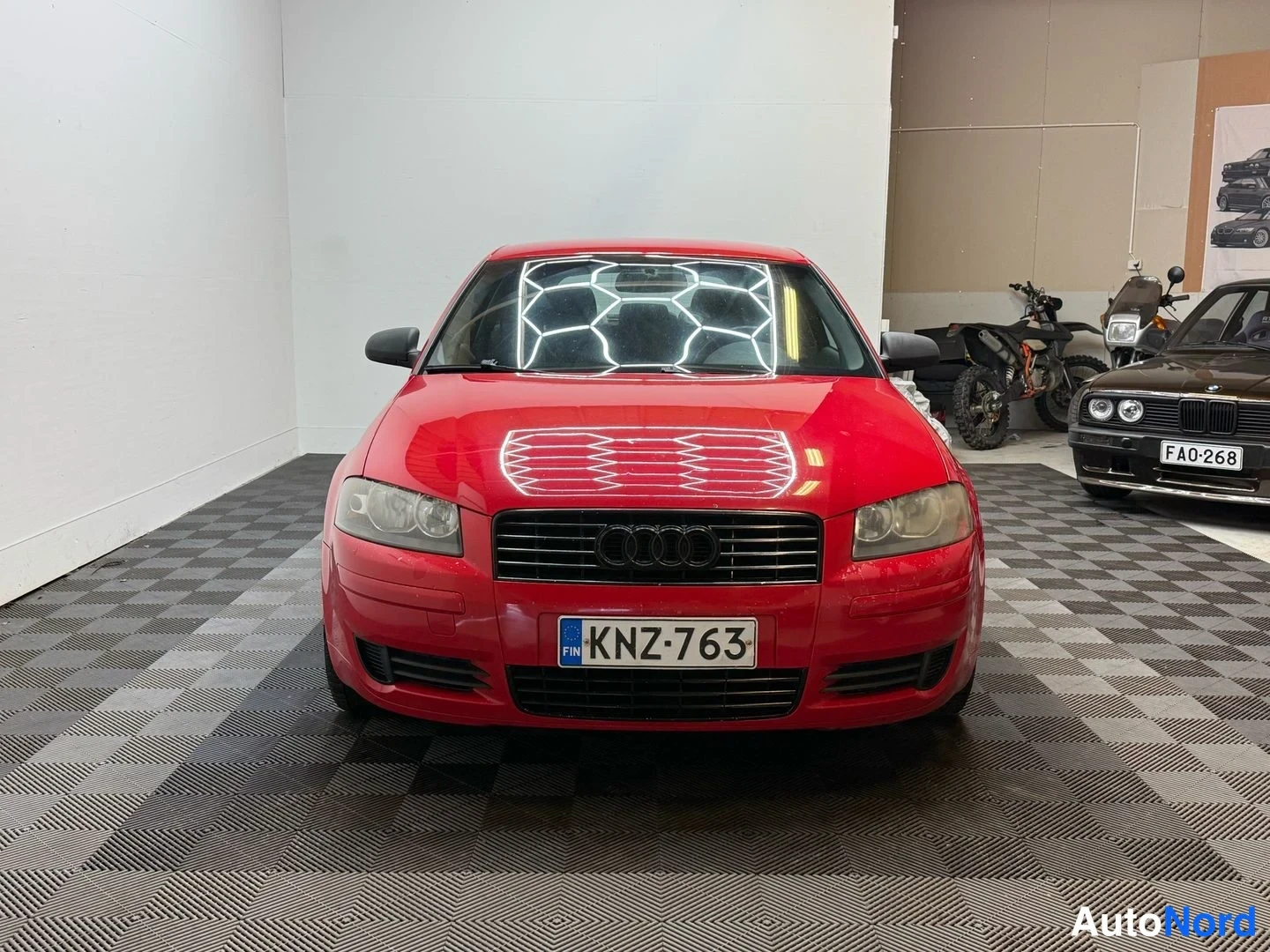 Edestä: Audi A3 Sportback, punainen, suora etunäkymä; etugrilli Audin-merkillä, etuvalot ja rekisterikilpi näkyvät hallissa.