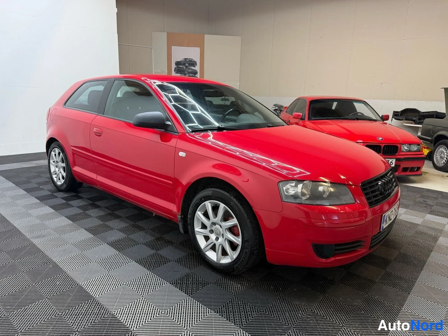 Etuviisto, nokka oikealle: Punainen Audi A3 -viistoperä kolmesta neljänneksestä edestä oikealta, taustalla punainen ja harmaa auto, sisätiloissa.