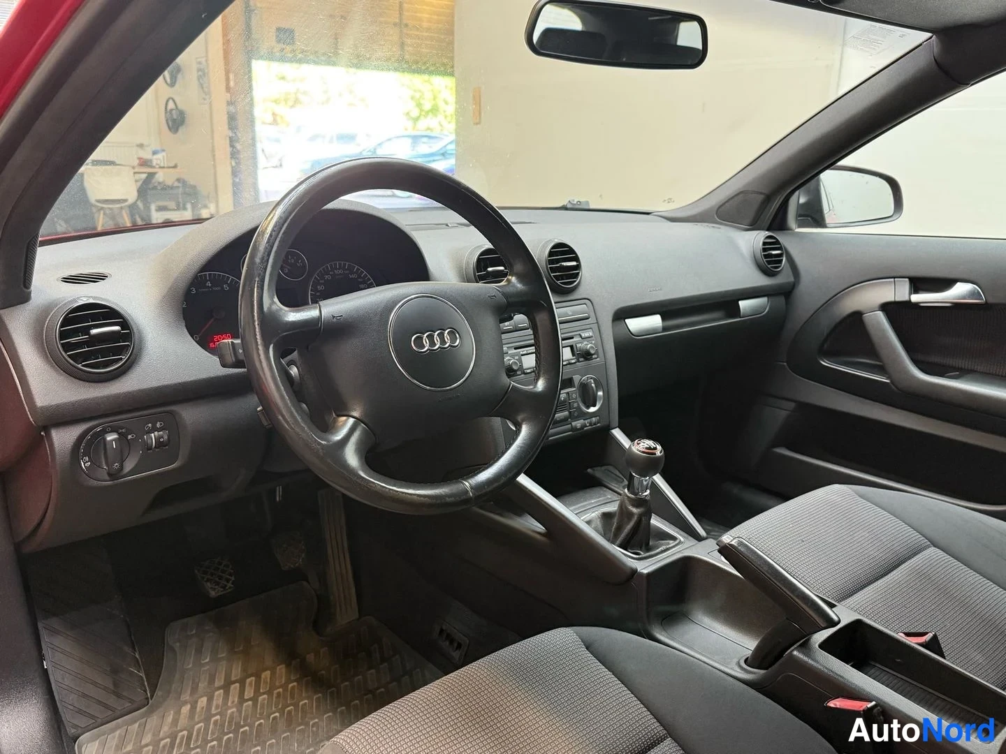 Muu: Ohjaamo tummalla kojelaudalla: Audi-merkillinen ohjauspyörä, mittaristo, käsivaihteisto ja radiokonsoli; kuljettajan ovi auki.