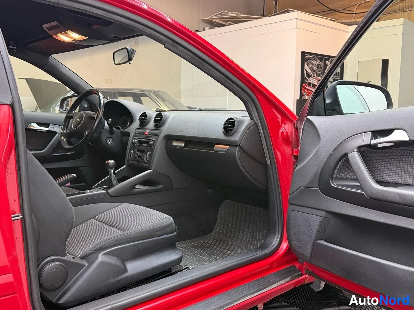 Muu: Audi A3:n sisätilat avoimen oven läpi: tumma sisusta, ohjauspyörä, manuaalivaihtehan ja kumimatot näkyvät.