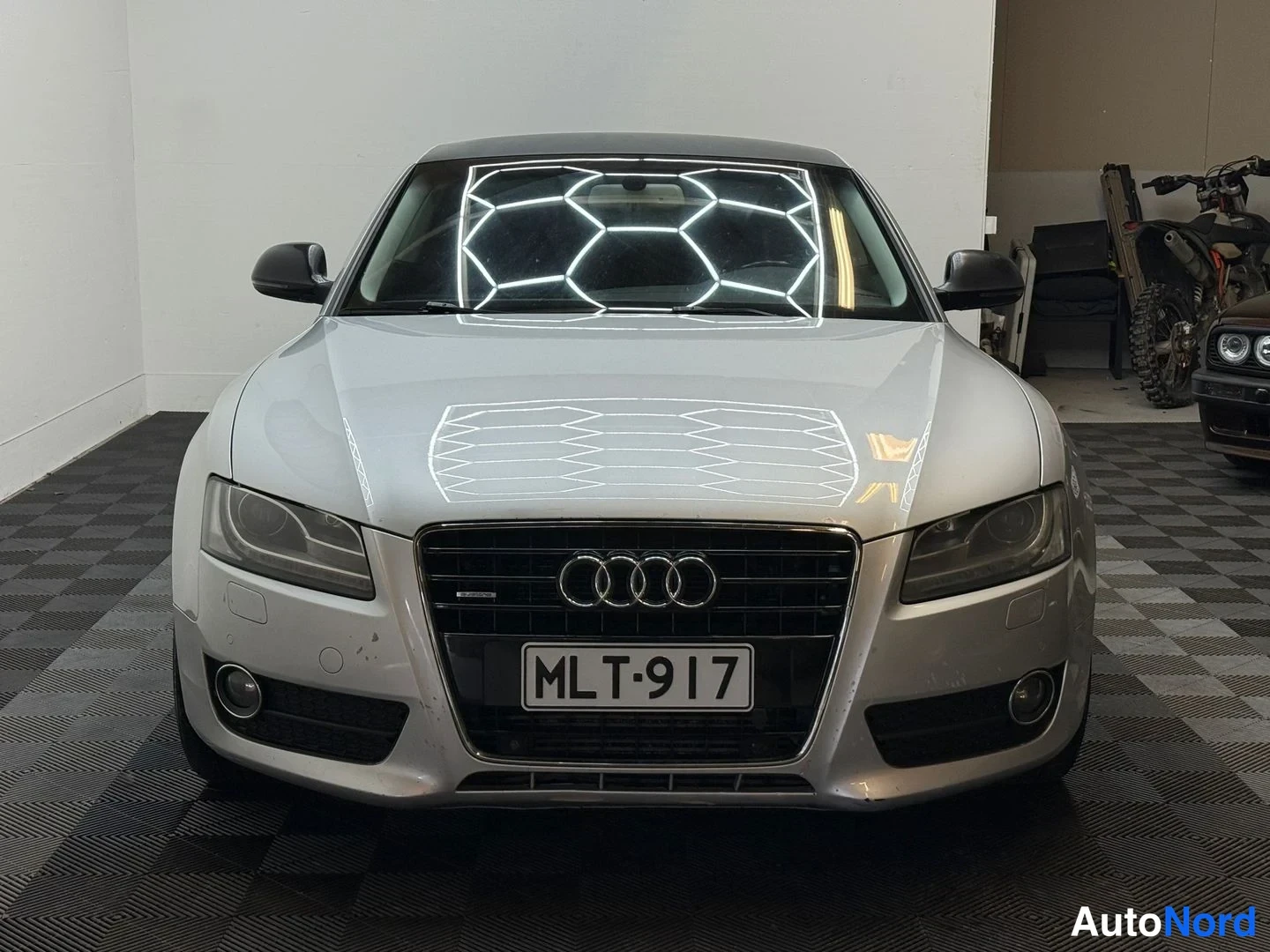 Edestä: Audi A5 -kupéHopeanvärinen henkilöauto sisätiloissa, suora etunäky, rekisteritunnus, keskellä näkyvä etusäleikkö.