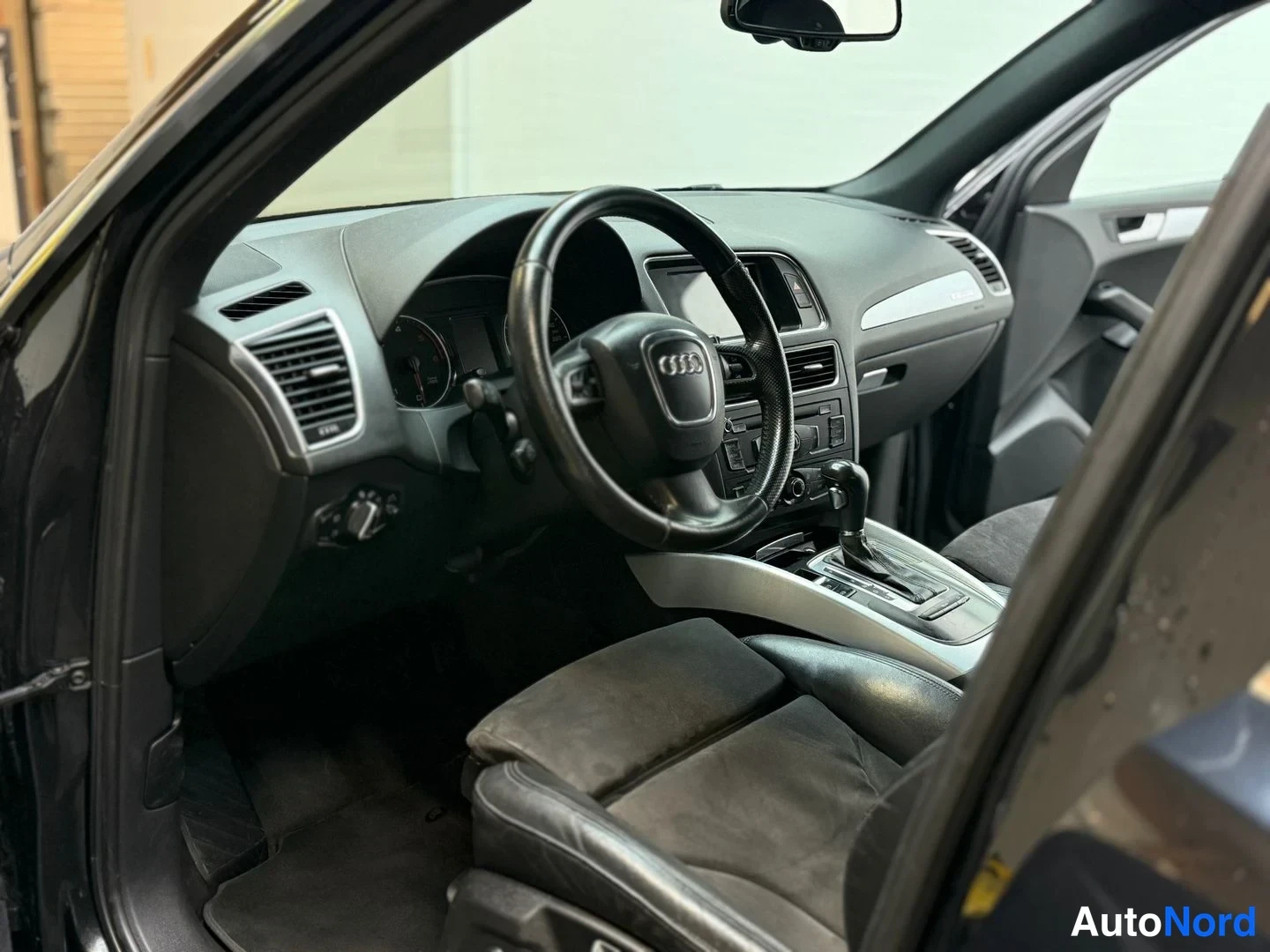 Muu: Audi Q5:n ohjaamo vasemmalta kuvattuna: Audi-merkkinen ohjauspyörä, mittaristo, keskikonsoli automaattivaihteen valitsimella ja tummat istuimet.
