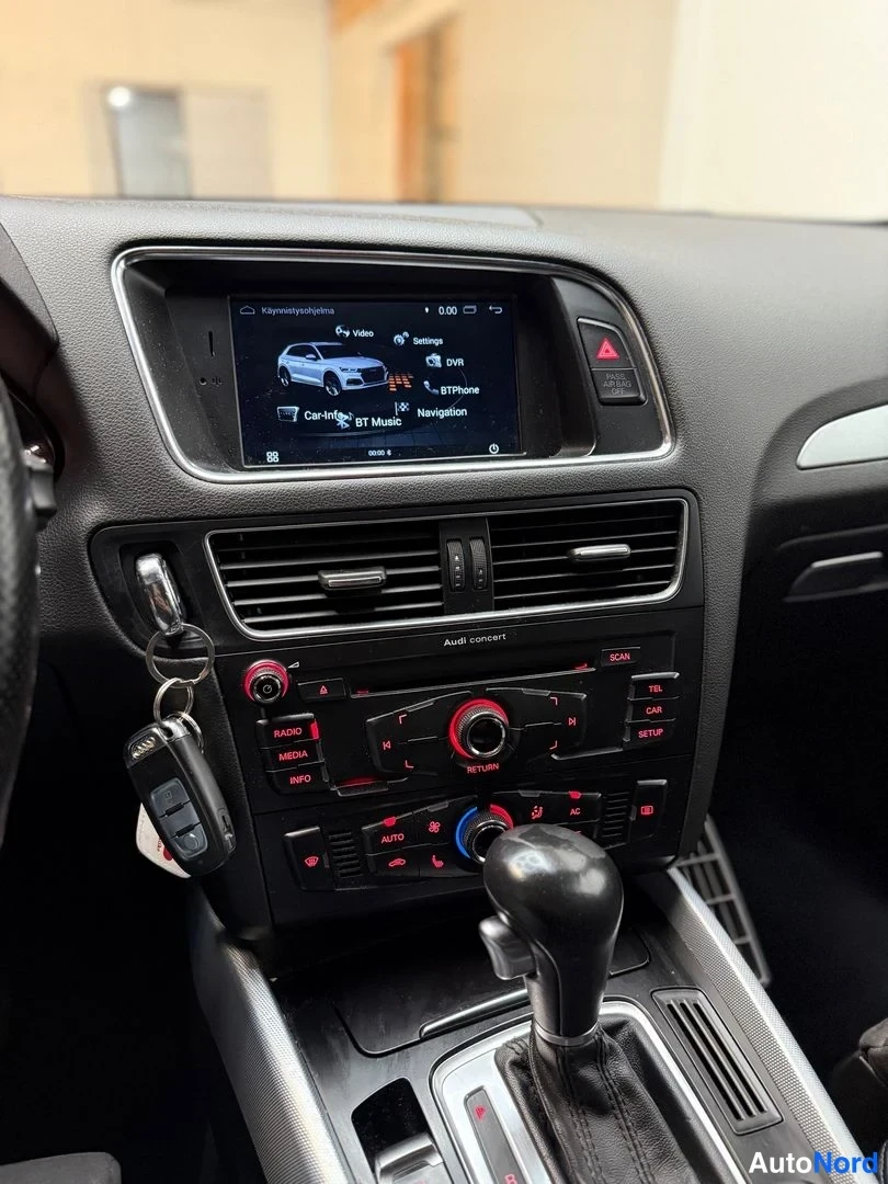 Sivulta: Kojelaudan keskiosan lähikuva: infotainment-näyttö, Audi concert -radion hallintapaneeli, ilmastointiautomaatin säätimet ja automaattivaihteen valitsin. Avaimet