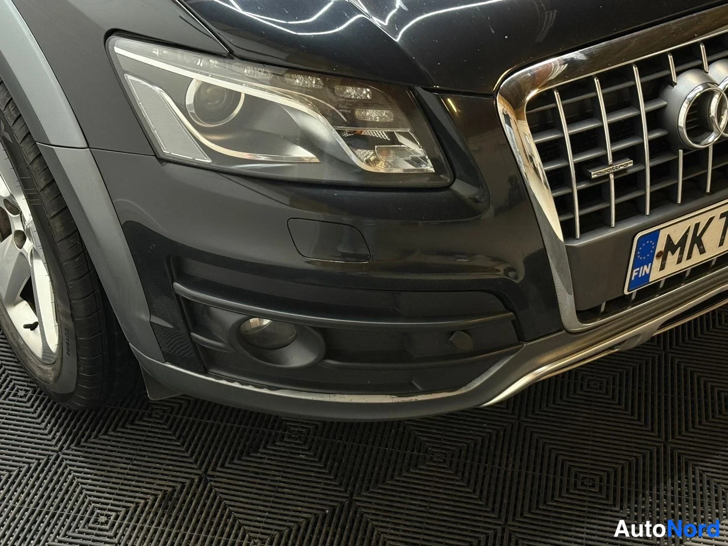 Edestä: Mustan Audi Q5:n etupuolen lähikuva: ajovalo, etusäleikkö ja etupuskuri rekisterillä MKT-... halliissa.