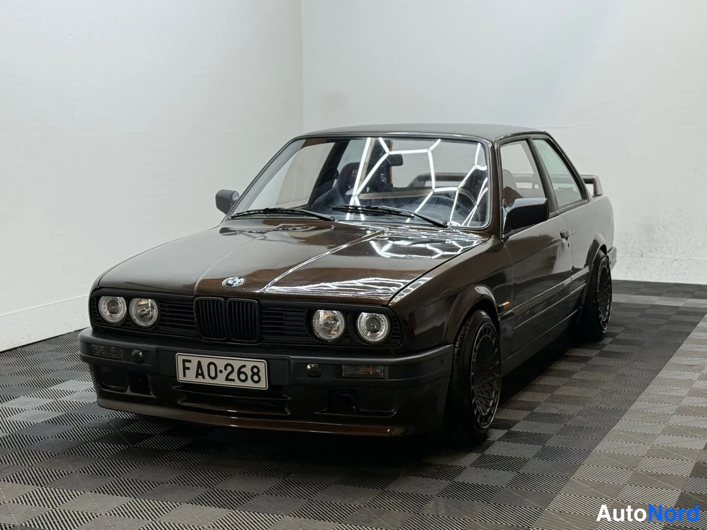 Etuviisto, nokka vasemmalle: Tummanruskea BMW-tyylinen kahden ovemallinen sedan kulmassa kuvattuna edestä vasemmalta; etumoitus, pyöreät ajovalot ja FA0-268-rekisterikilpi näkyvät.