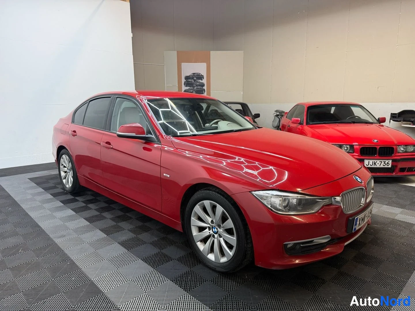 Etuviisto, nokka oikealle: Punaisen BMW-sedanin etukulmanurkku varastotilassa, etumoottoritila ja oikea kylki näkyvät; rekisterikilpi ja muut ajoneuvot taustalla.